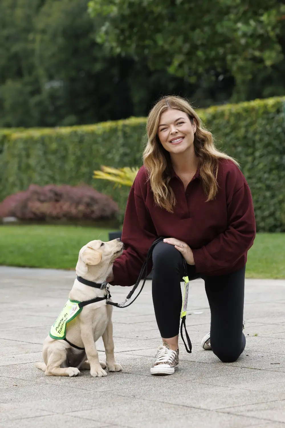 Woman kneeling and smiling next to a sitting Labrador puppy wearing a green training vest in a park setting.