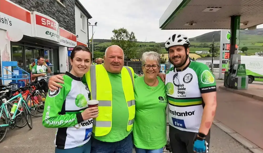 Four people wearing green Irish Guide Dogs charity gear pose together at a gas station during a cycling event, with bicycles and rural landscape visible in the background.