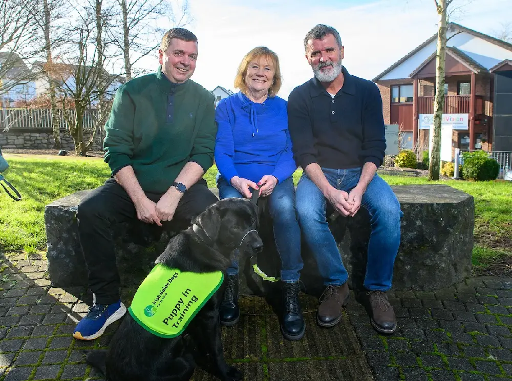 Three adults sit on a stone bench with a black service dog wearing a green "Puppy in Training" vest in a residential garden setting.