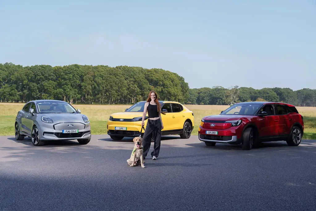 Roz Purcell standing holding the lead of an IGD dog with 3 Ford cars in the background