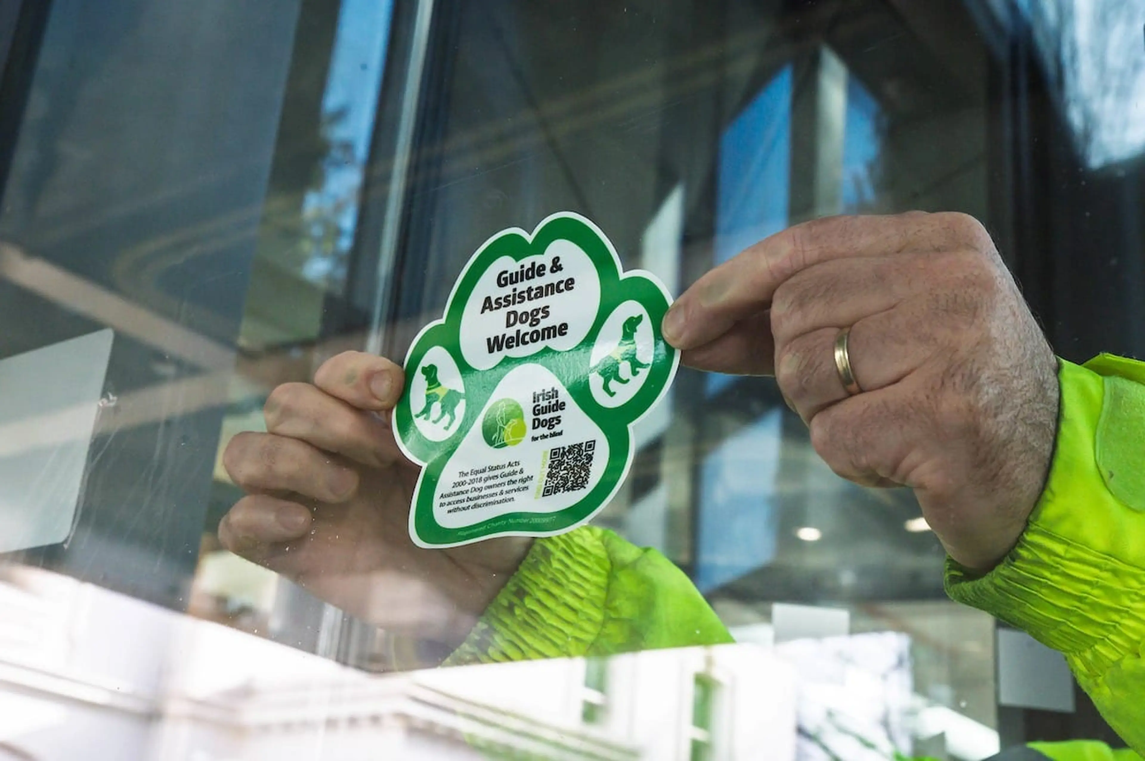 A person in a lime green jacket holds up a green paw-shaped sticker that reads "Guide & Assistance Dogs Welcome" with the Irish Guide Dogs logo and a QR code.