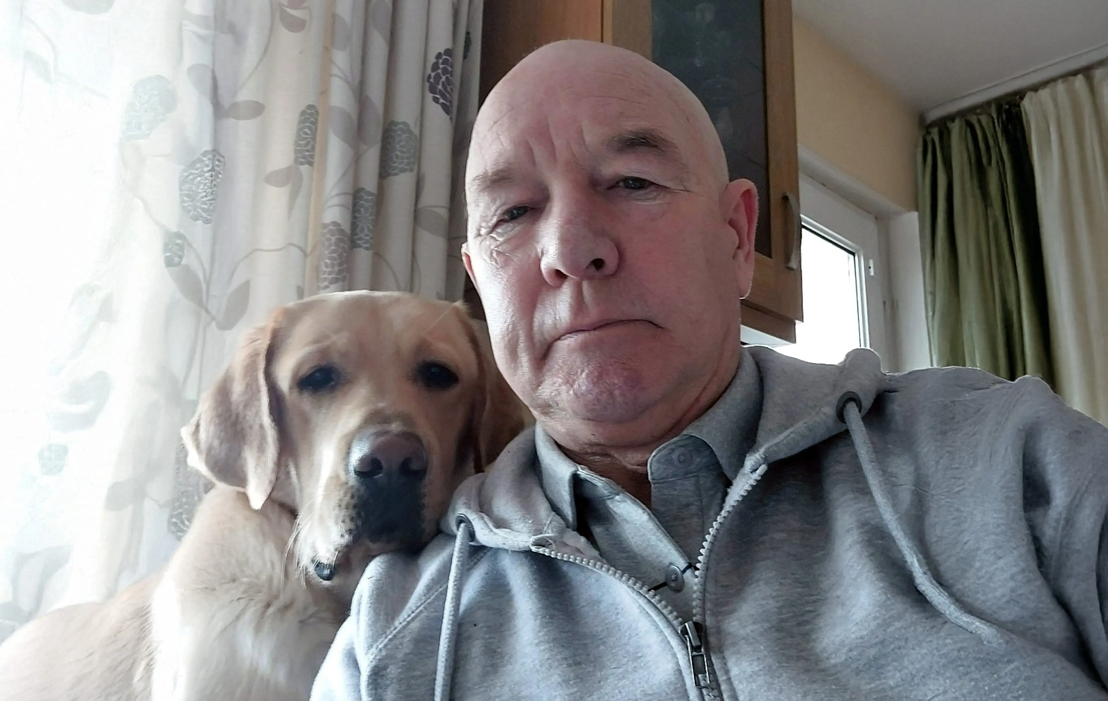 A middle-aged man in a gray jacket poses for a selfie indoors with a light-colored Labrador Retriever beside him, near a window with patterned curtains.