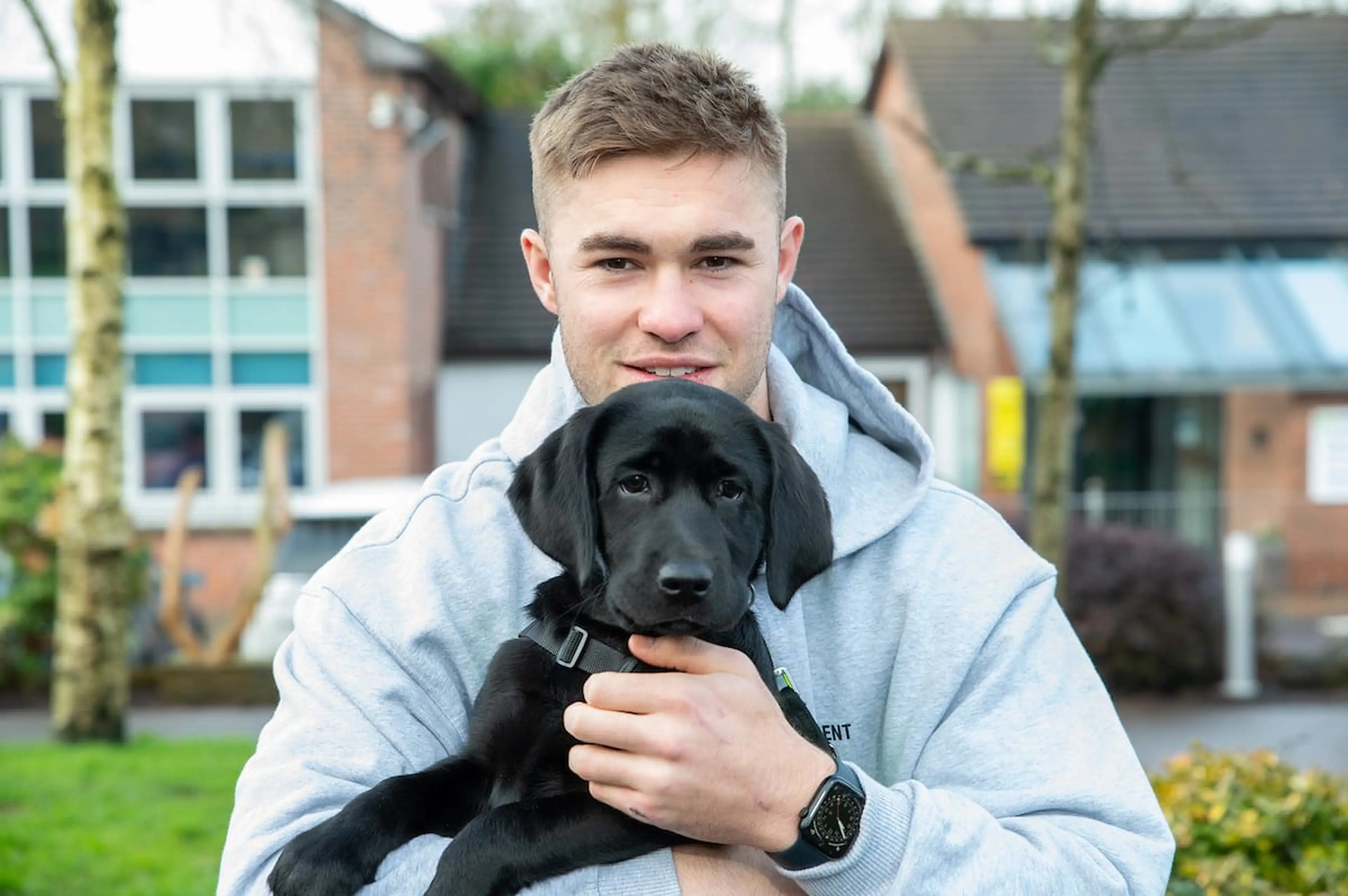 A smiling young man wearing a gray hoodie is holding a cute black Labrador puppy in front of a building.