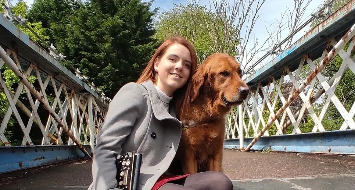 A woman in a gray coat kneels on a bridge, smiling, next to a golden retriever. Lush greenery surrounds the background.