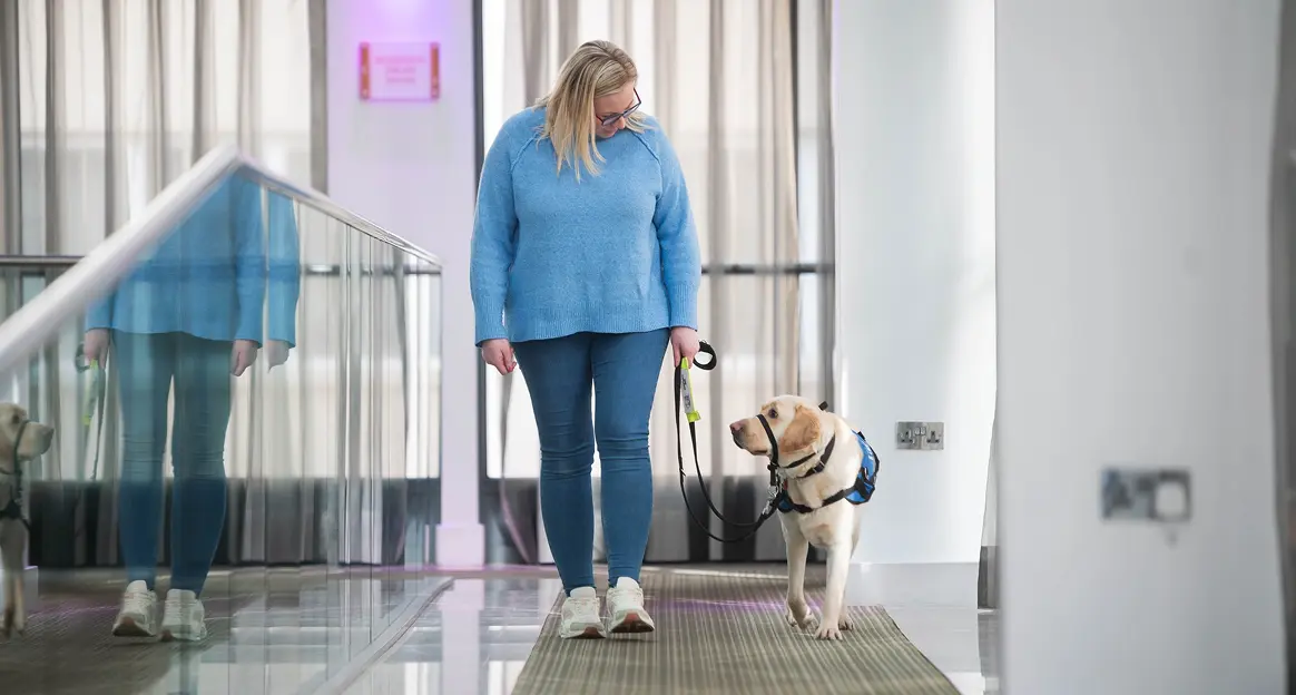 A woman in a blue sweater walks with a yellow Labrador service dog on a leash through a modern, bright hallway with glass railings.