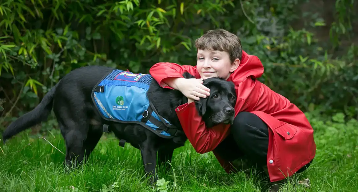 A child in a red coat hugs a black dog wearing a blue vest in a grassy area with bushes in the background.