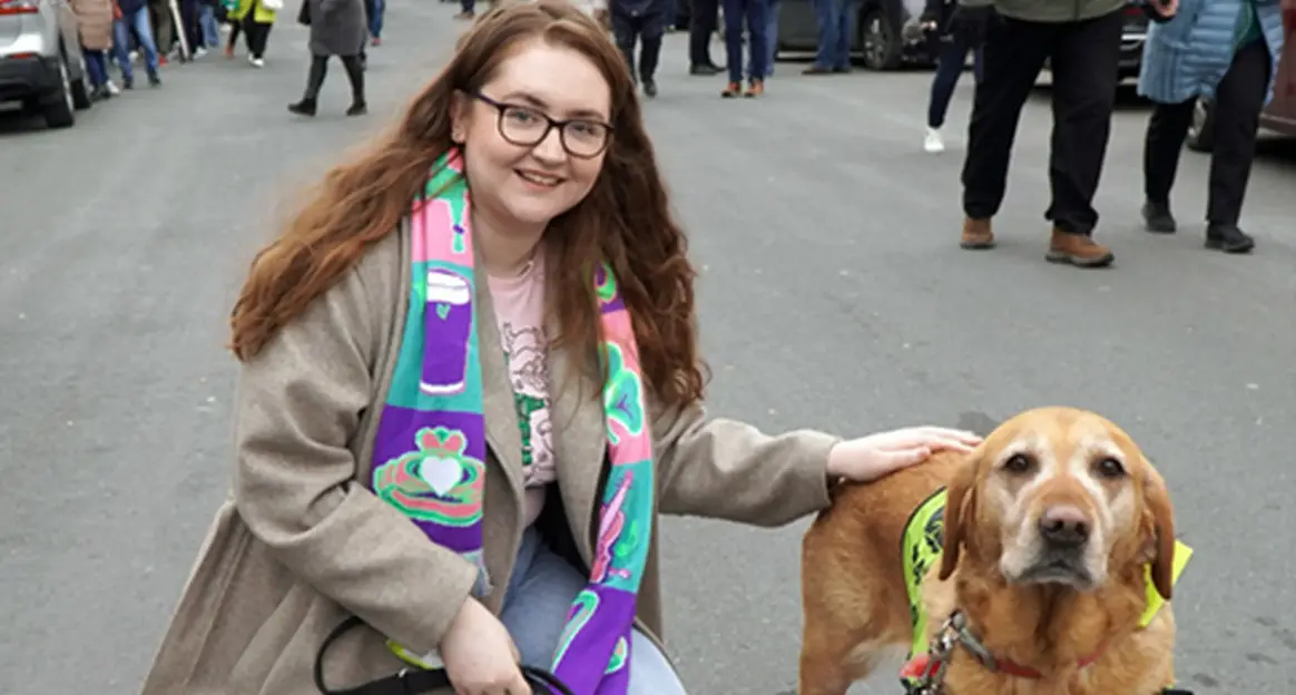 A smiling young woman with long reddish-brown hair and glasses wearing a beige cardigan and colorful scarf stands on a city street while petting a brown service dog wearing a yello