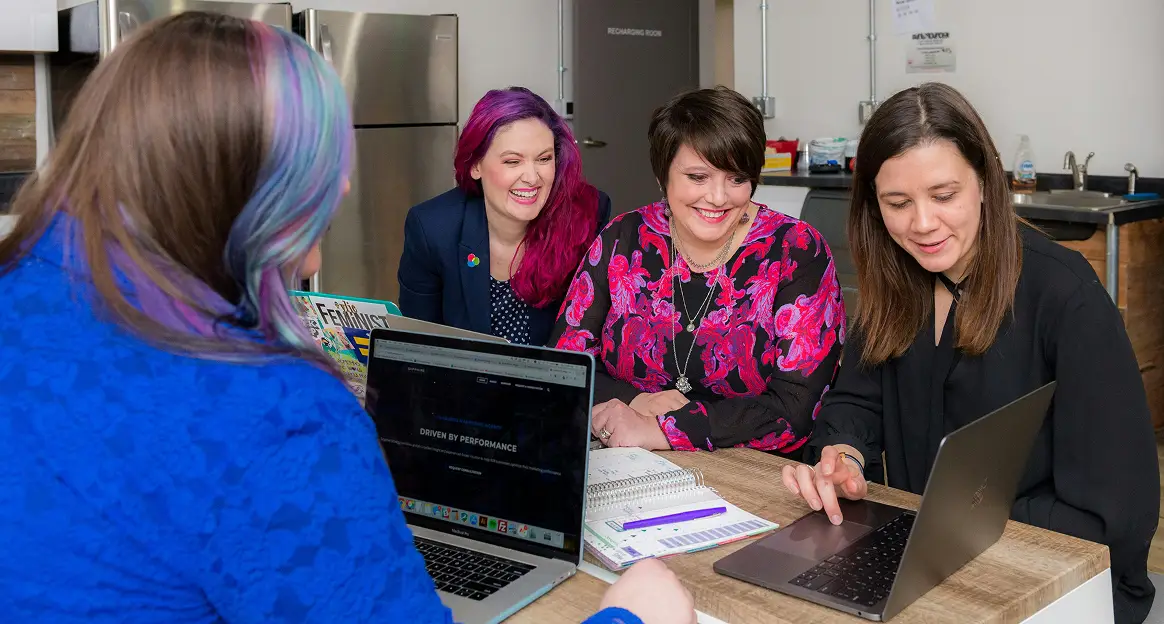 Four women collaborate at a table with laptops and a planner, smiling and discussing a project in a modern office setting.