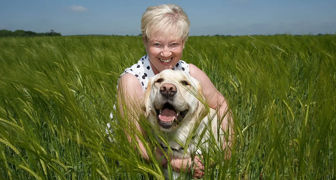Smiling woman hugging a happy Labrador retriever in a lush green field under a clear blue sky.