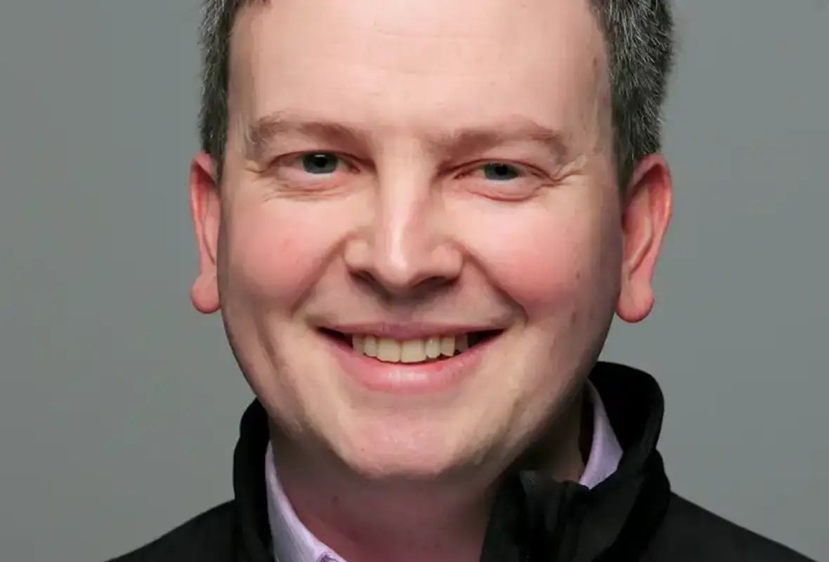 A man with dark hair and a warm smile wearing a black jacket over a light purple shirt, photographed against a neutral gray background.