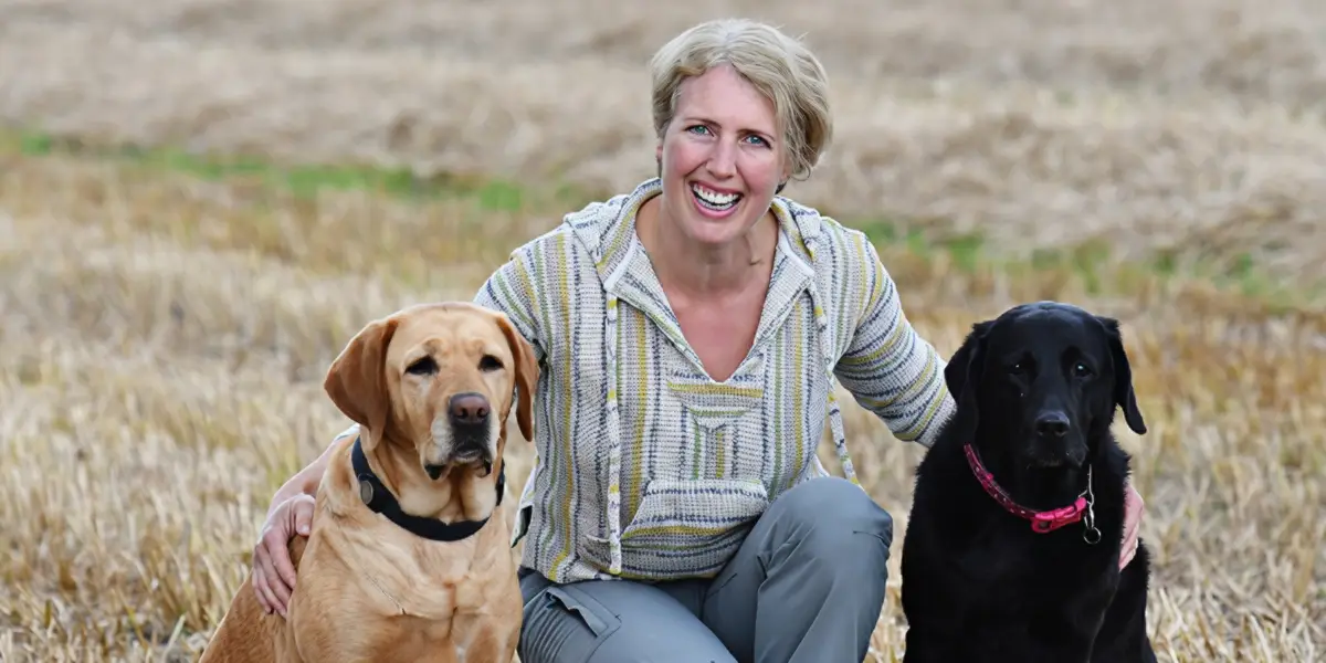 A smiling woman with blonde hair crouches in a harvested field while posing with a brown Labrador on her left and a black Labrador wearing a pink collar on her right.