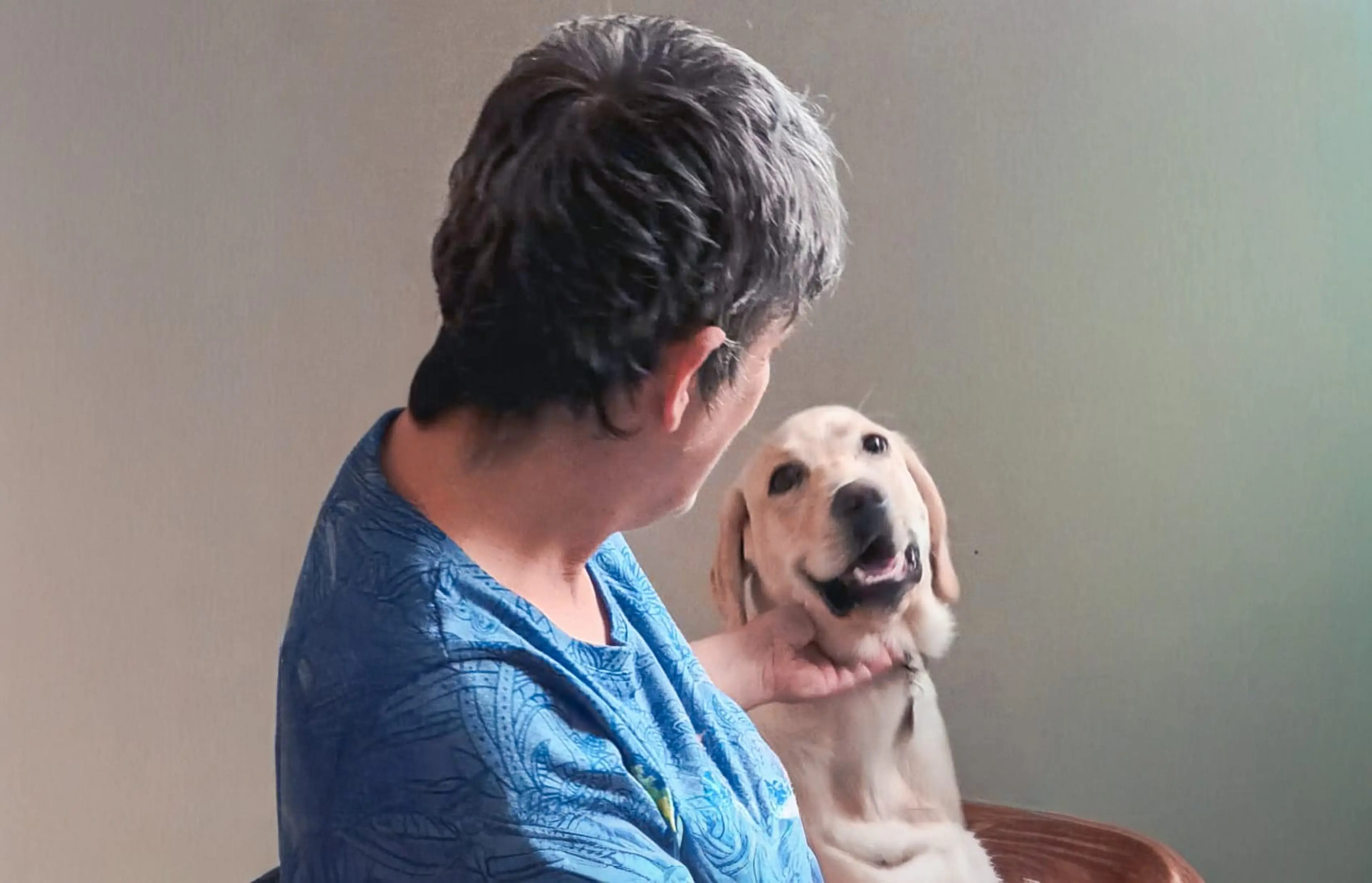 A person with short dark hair wearing a blue patterned shirt holds a light-colored Labrador Retriever that appears to be smiling at the camera.