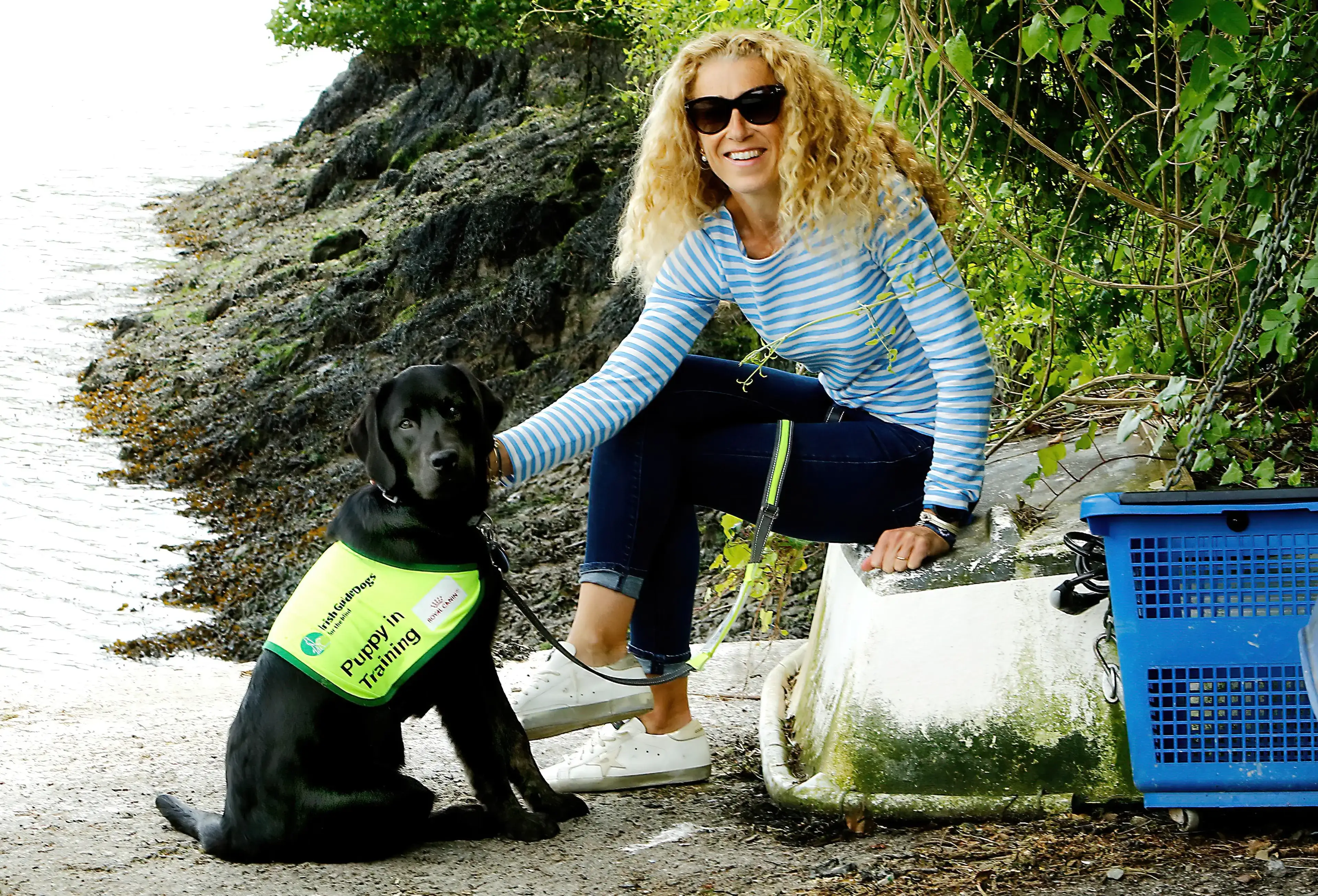 A young woman with curly blonde hair and sunglasses sits next to a black Labrador retriever wearing a "Puppy in Training" vest, both smiling in a natural outdoor setting.