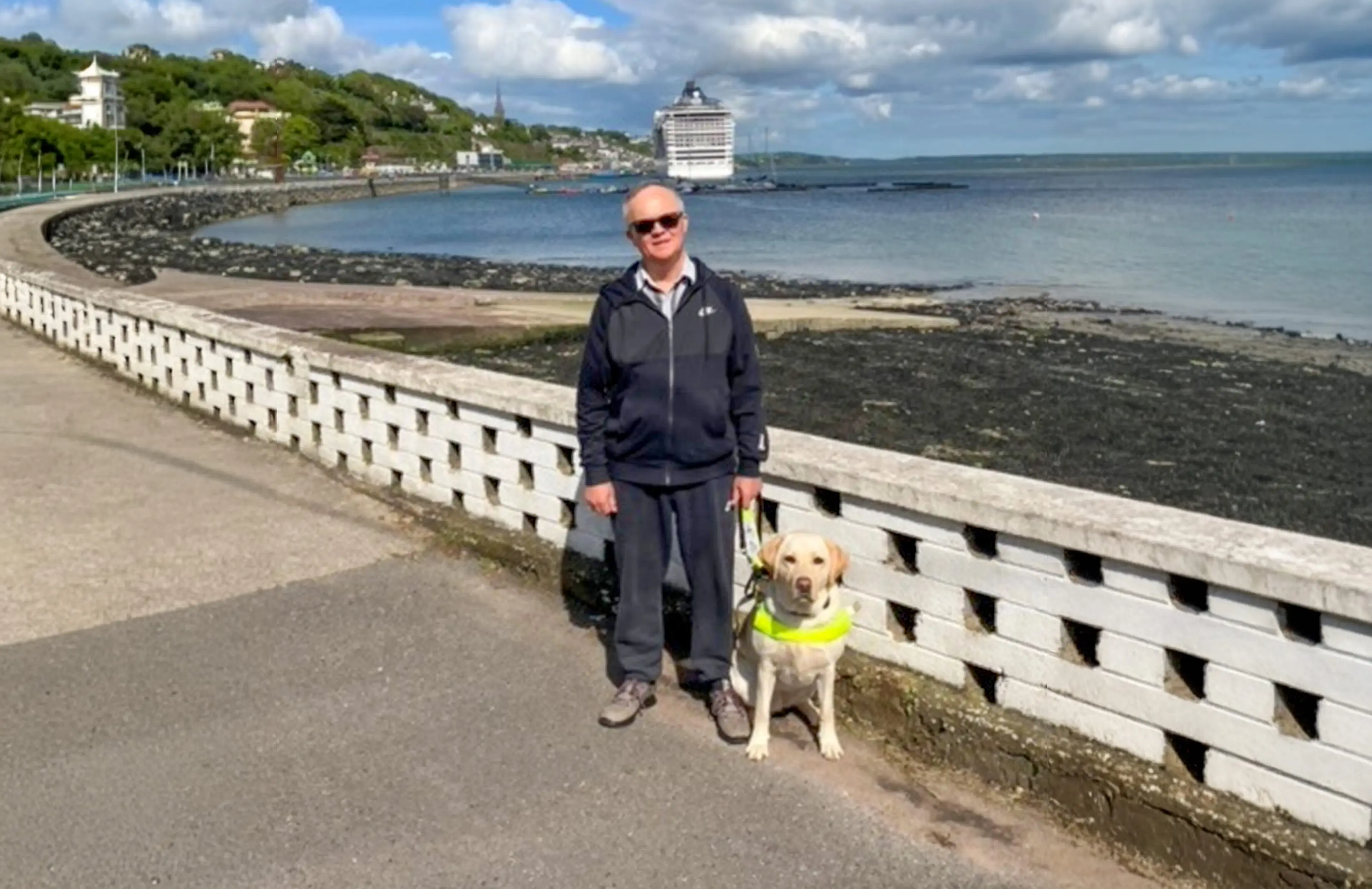 A man wearing sunglasses and a dark jacket stands with a yellow Labrador Retriever on a coastal promenade, with a large cruise ship and seaside town visible in the background.