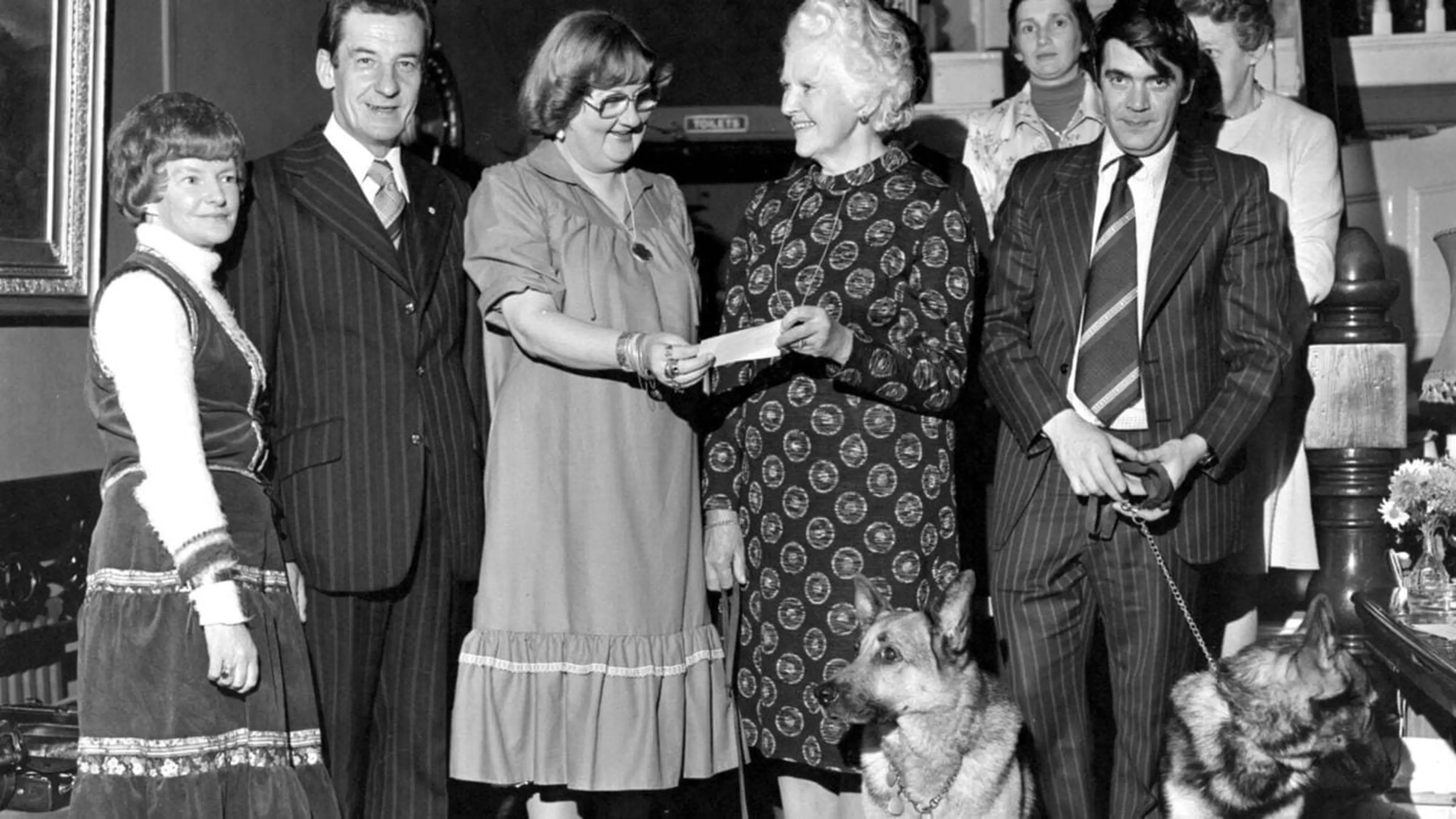 This black and white image shows a group of people, including a man and woman in formal attire, standing together and shaking hands in what appears to be a social or formal event, 