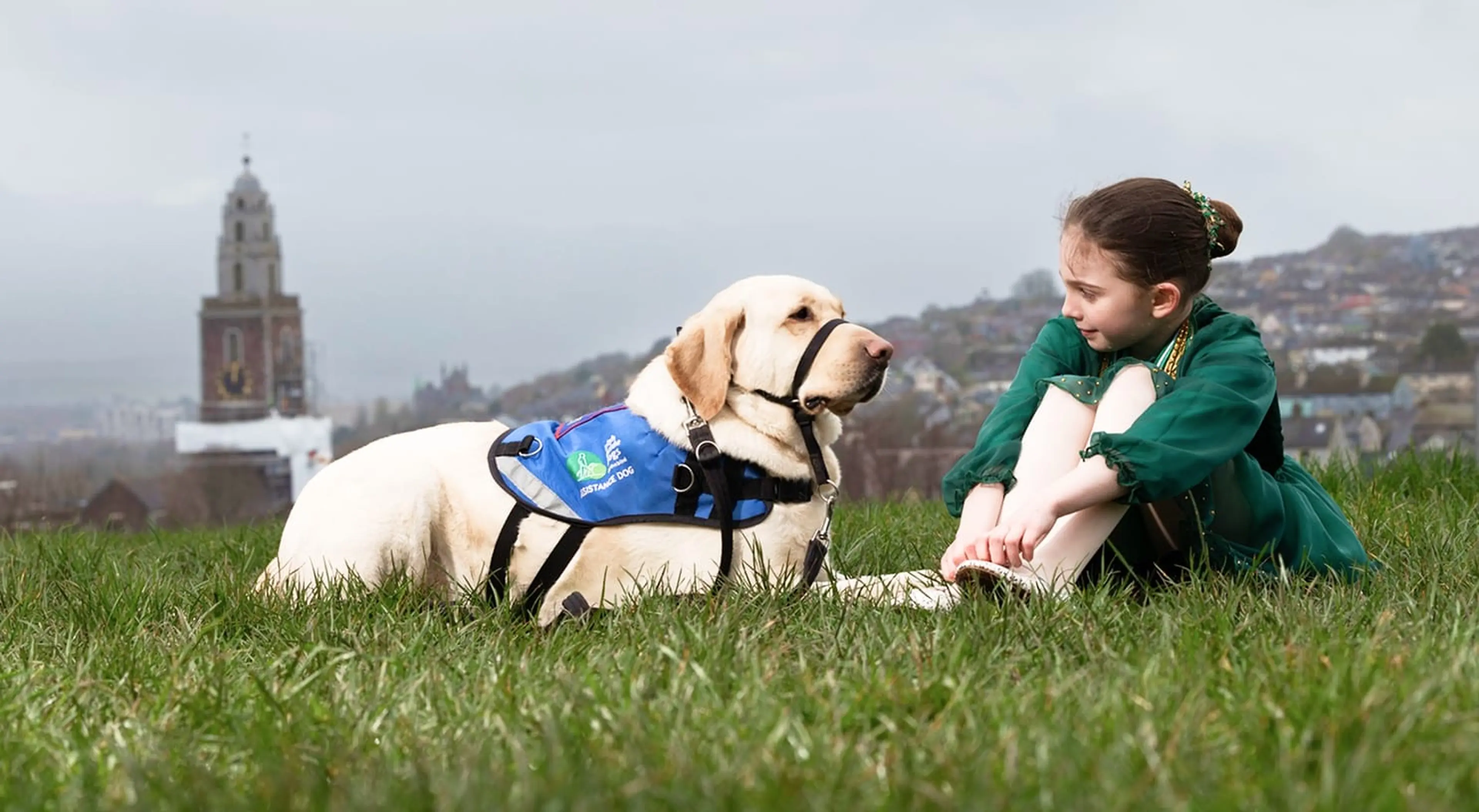 A young girl in a green jacket sits with a cream-colored service dog wearing a blue vest on a grassy hillside, with a church tower and residential buildings visible in the misty ba