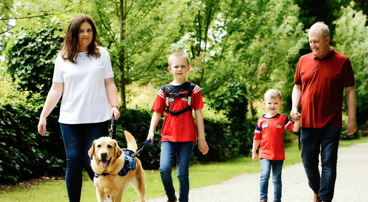A family of five walks together on a tree-lined path with a golden retriever service dog on a leash.
