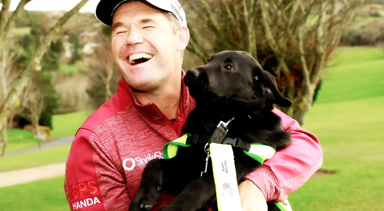 A man in a red SPS Honda polo shirt and white cap smiles while holding a black dog with a green and white harness in a garden setting.