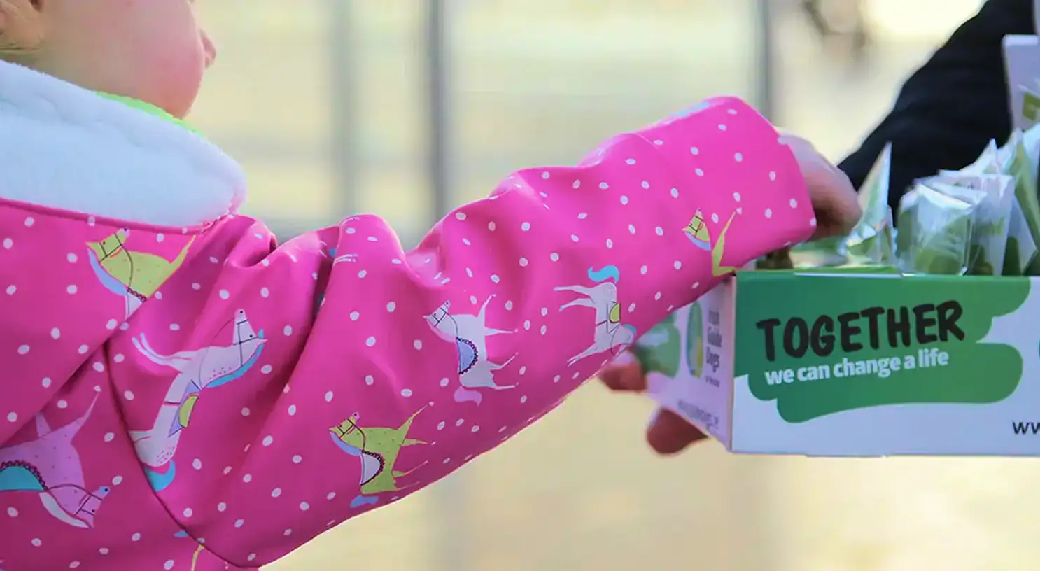 A child wearing bright pink mittens with white polka dots and playful animal designs holds a green box labeled "TOGETHER we can change a life" during what appears to be a charitabl