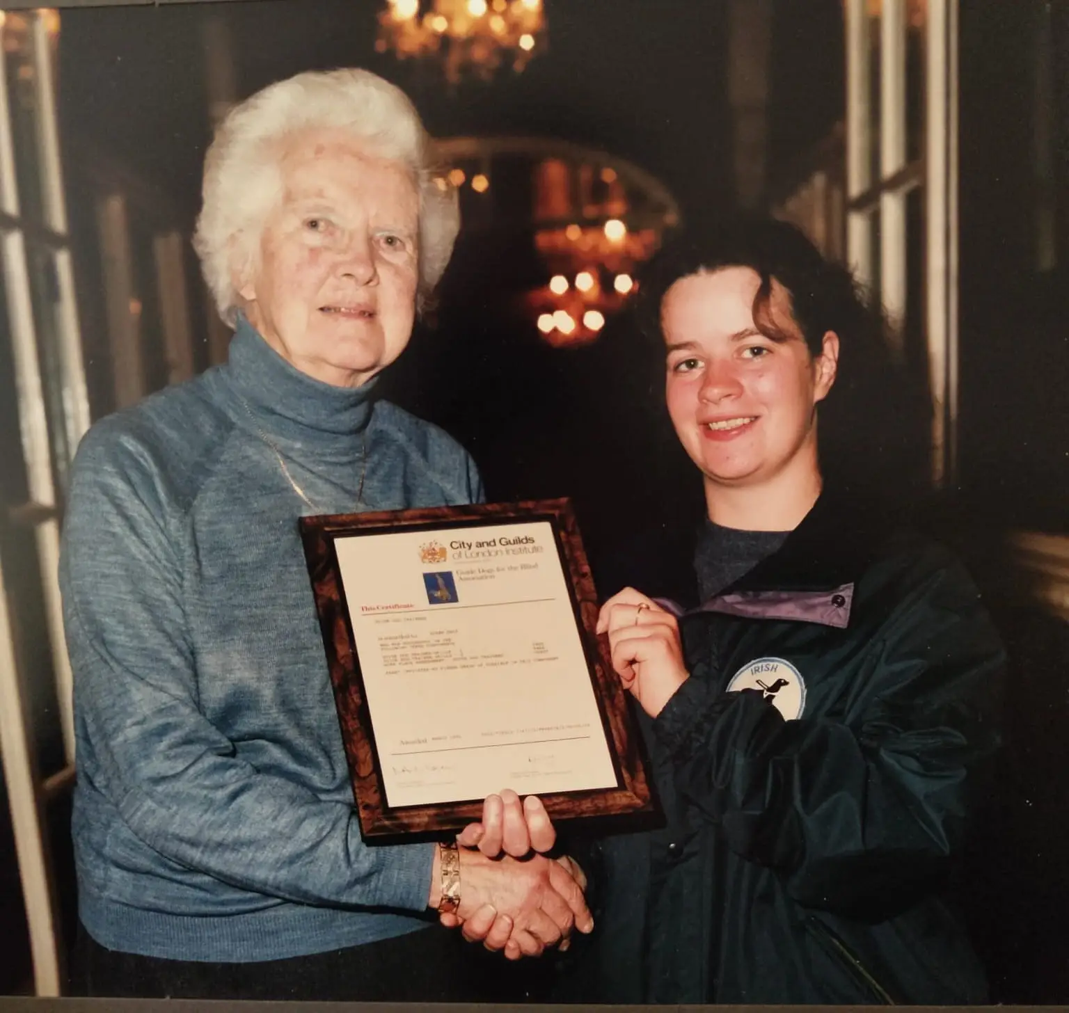 Two women smiling, one elderly and one young, holding a framed certificate in a warmly lit room with chandeliers.