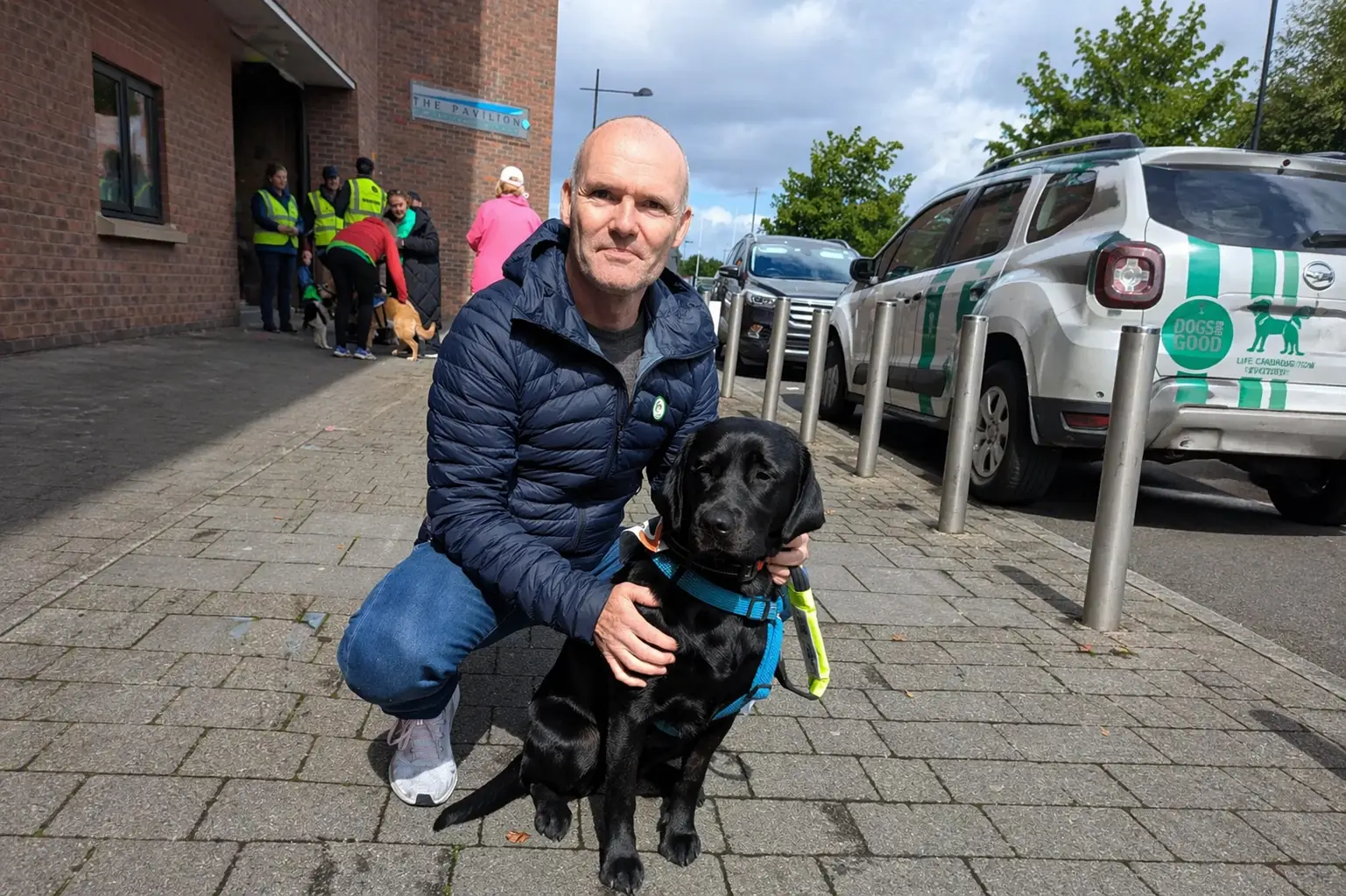A man in a dark blue puffy jacket crouches beside a black Labrador wearing a blue harness outside a brick pavilion building, with a green and white van and other people visible in 