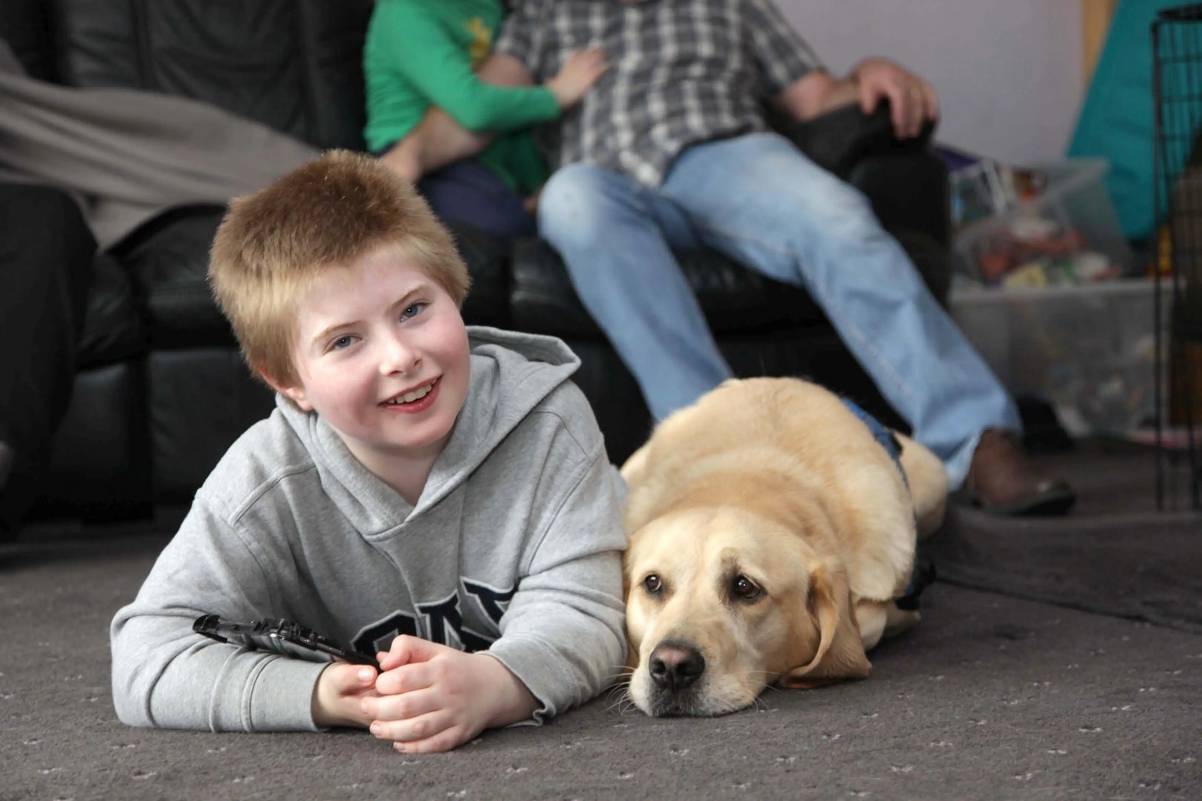 A young boy in a gray hoodie lies on the ground smiling while petting a golden Labrador retriever, with other children sitting nearby.