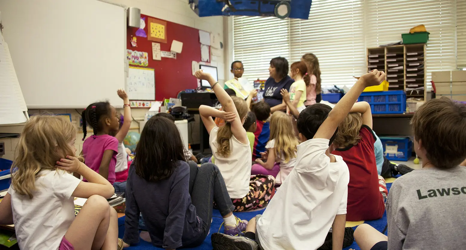 A diverse group of elementary school students sit on a blue carpet in a classroom while raising their hands during a lesson led by a teacher and teaching assistants.