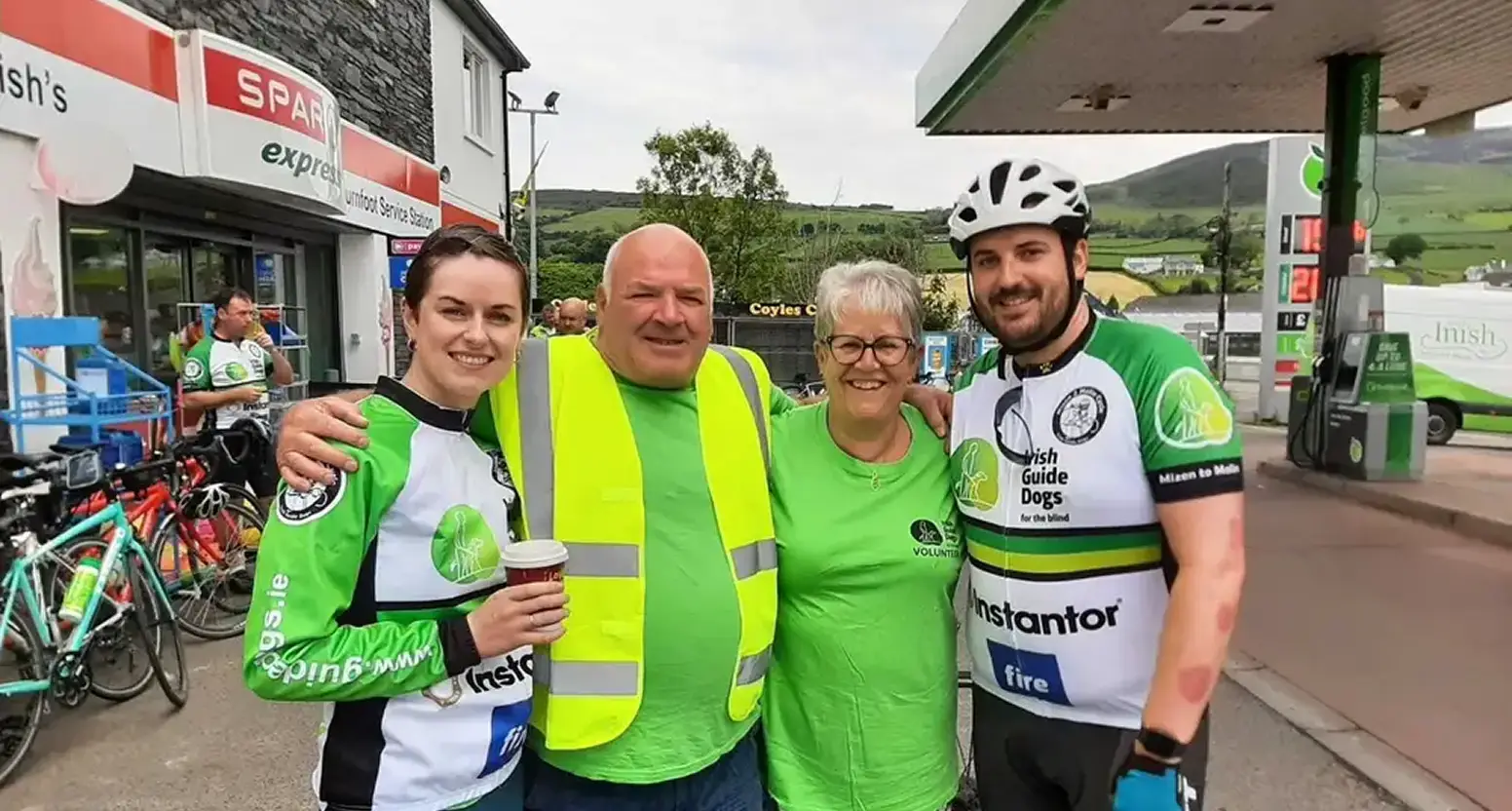 Four people in green athletic wear and safety vests pose together at a gas station during what appears to be a charity cycling event supporting Irish Guide Dogs for the Blind.