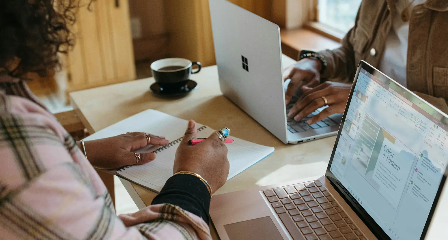 Two people collaborate at a wooden table with multiple laptops and a notebook, with a cup of coffee nearby in a bright, natural workspace.