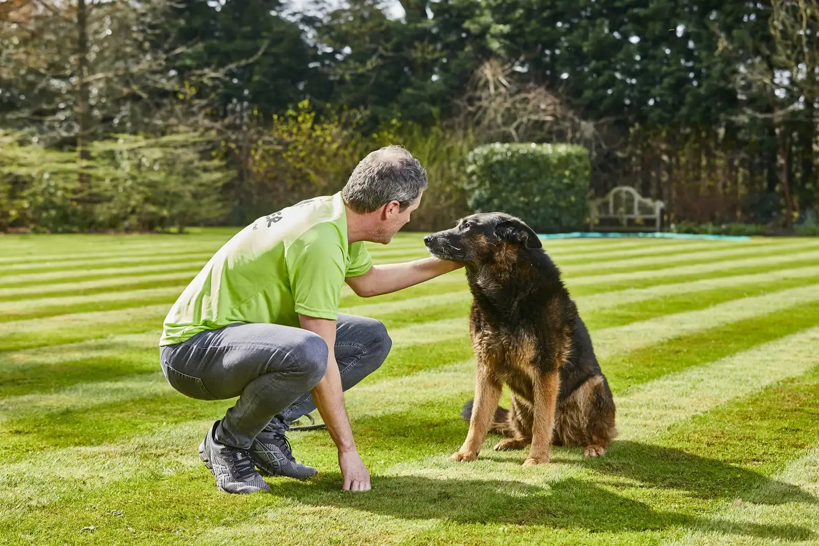 A man in a lime green shirt crouches down to pet a dark-colored dog sitting on a manicured lawn in a spacious garden.