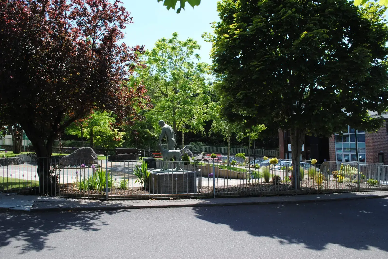 A public park featuring a central statue of a man with a horse, surrounded by a metal fence, flowering plants, and mature trees, with a brick building visible in the background.