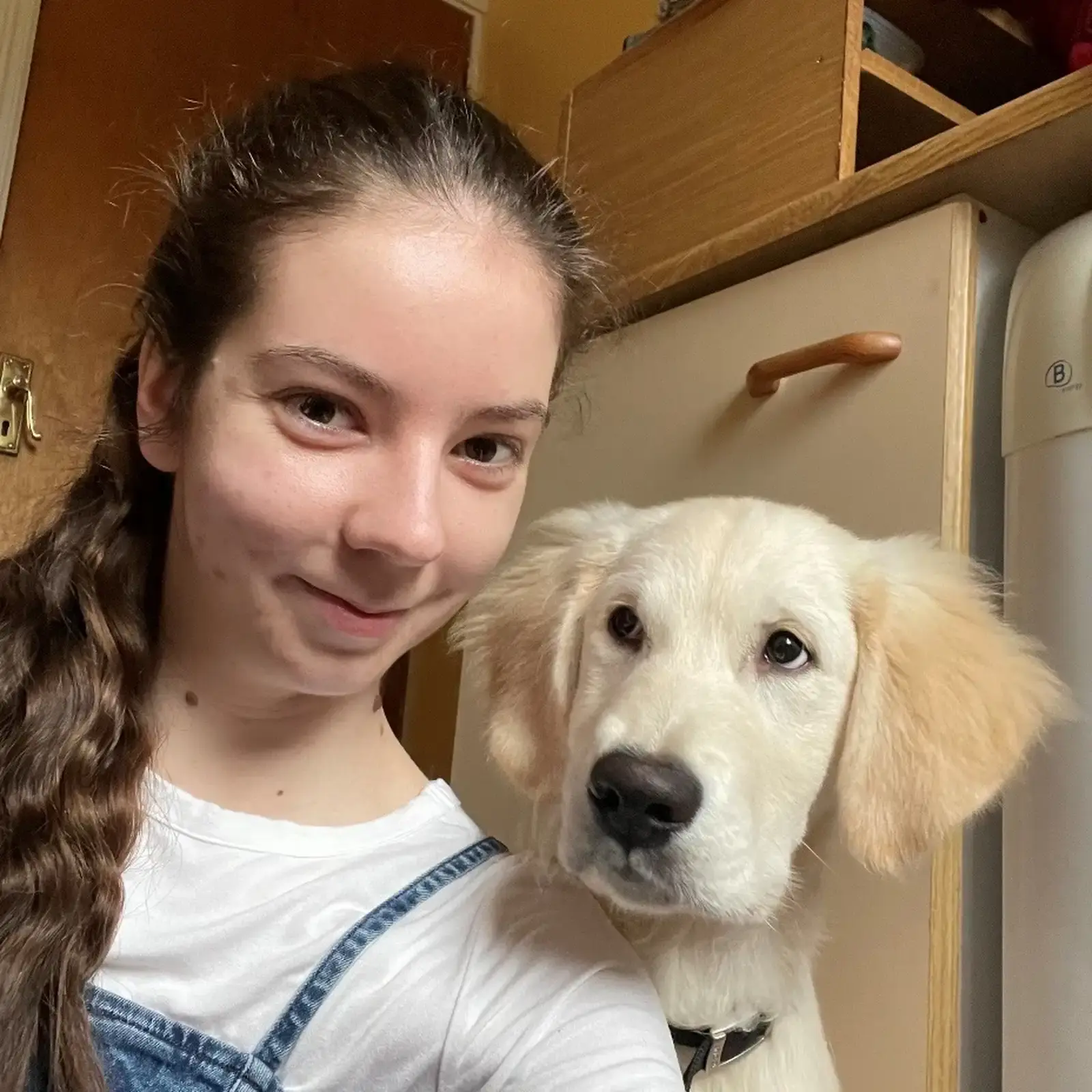 A young woman with long brown hair smiles while posing next to a cream-colored Labrador Retriever wearing a collar indoors.
