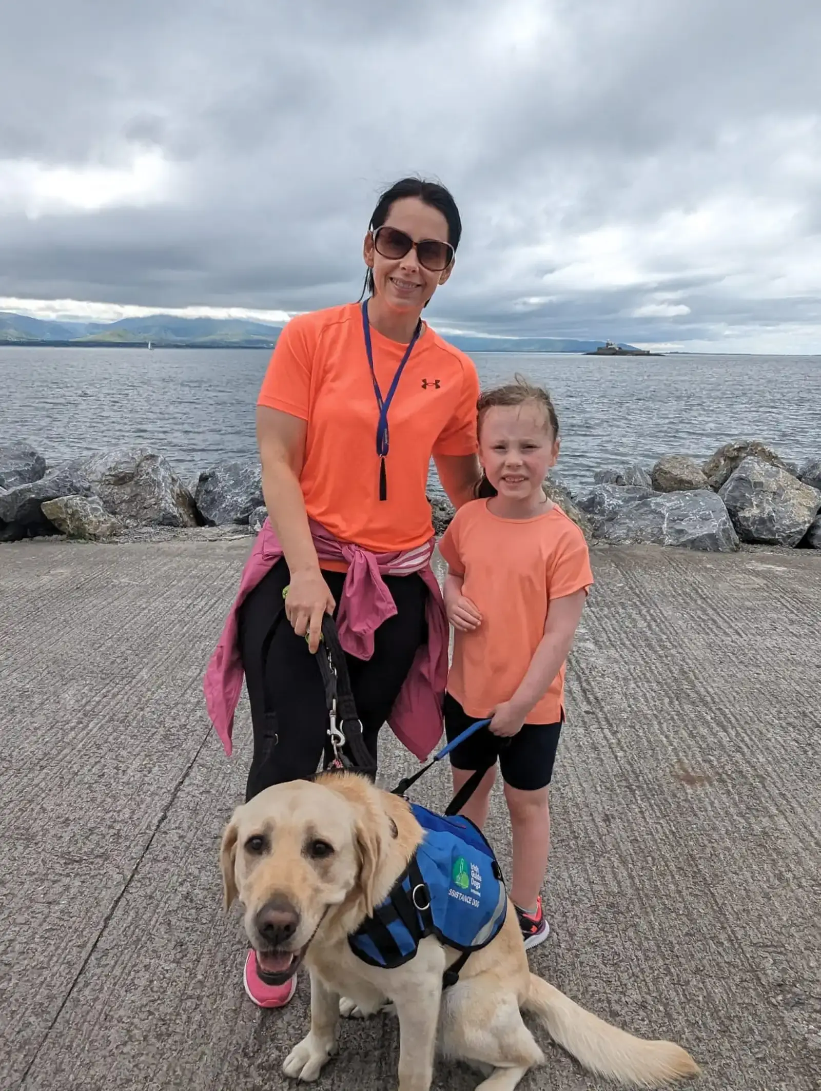 A woman and young child in orange shirts pose on a beach with a service dog wearing a blue vest, with mountains and cloudy skies visible across the water behind them.