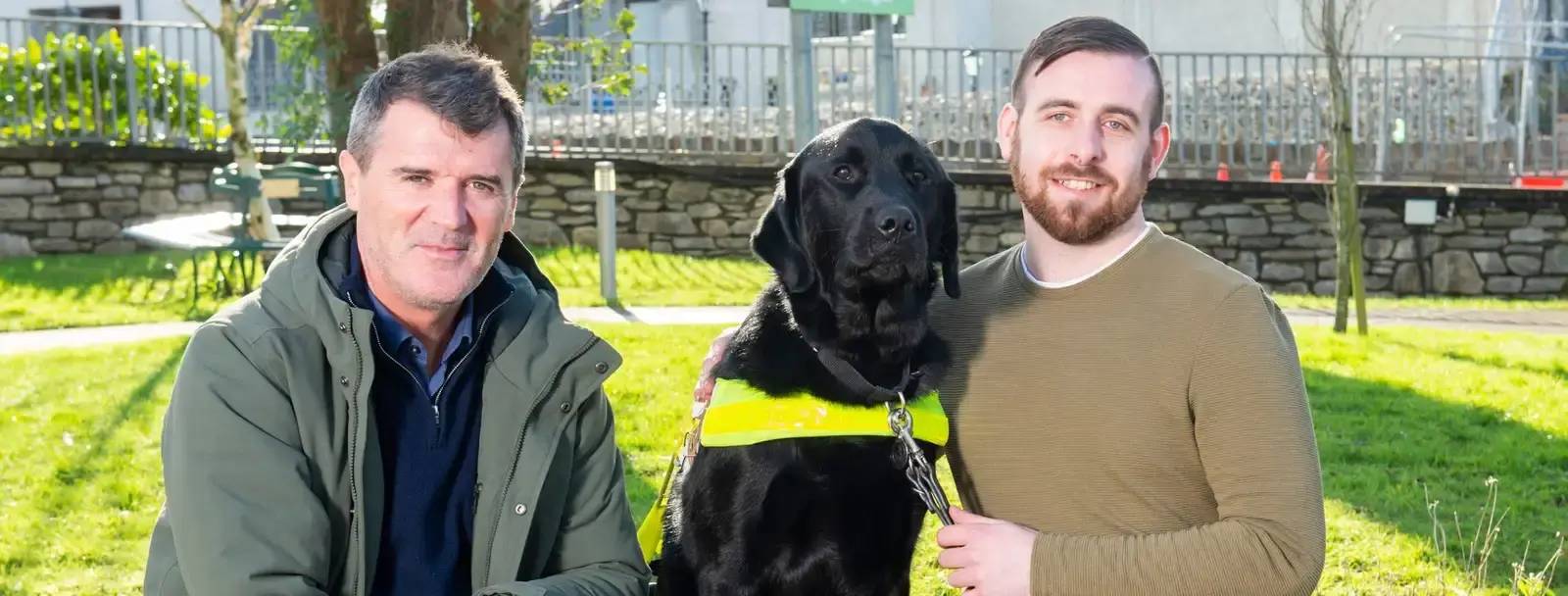 Two men pose with a black Labrador Retriever in a sunny park setting with stone walls and green grass in the background.