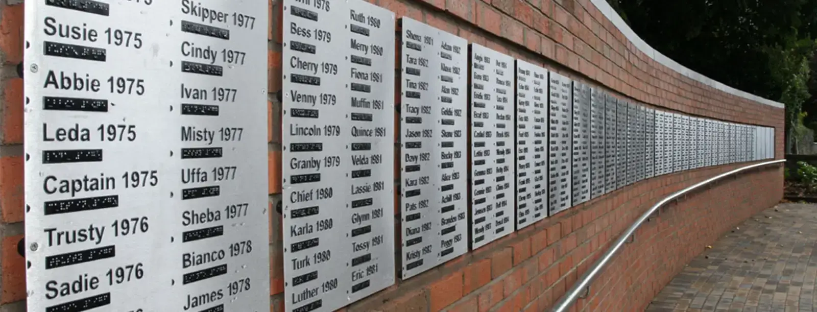 A brick memorial wall displays white plaques with names and years in chronological order from 1975 onwards, each with Braille translations beneath the text.