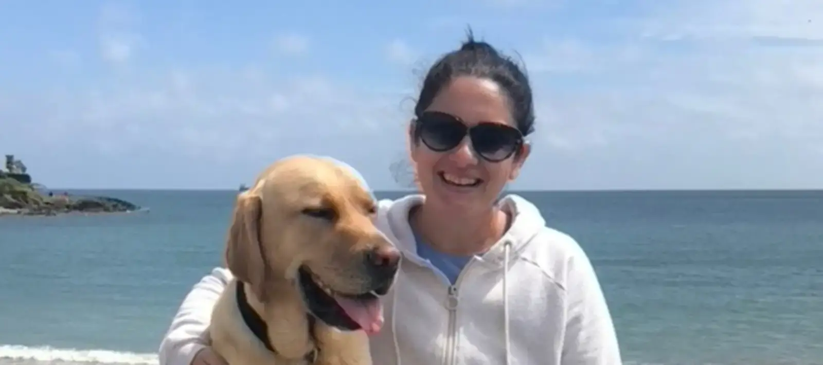A woman wearing sunglasses and a white hoodie smiles while posing with a yellow Labrador retriever on a sunny beach with turquoise water and rocky cliffs in the background.