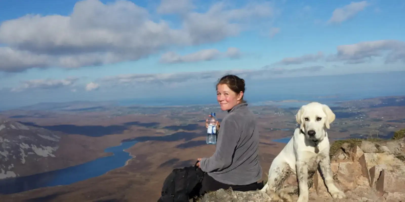 A woman and a white Labrador sit on a rocky mountain summit overlooking a vast landscape of forests, lakes, and valleys under a partly cloudy sky.