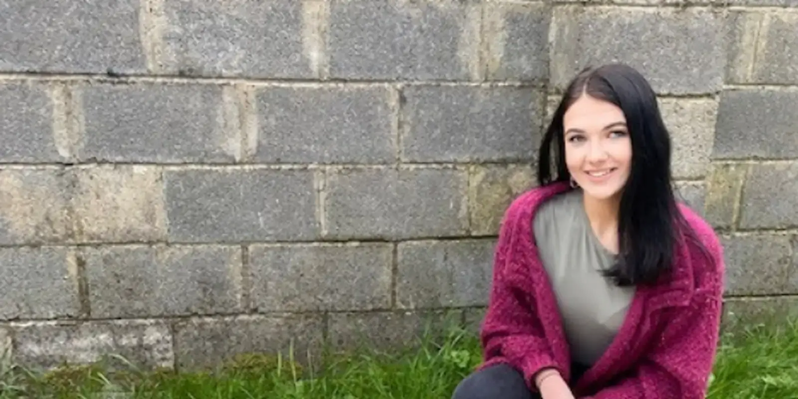 A young woman with long dark hair wearing a maroon cardigan over a gray top sits smiling against a gray stone brick wall with grass at the base.