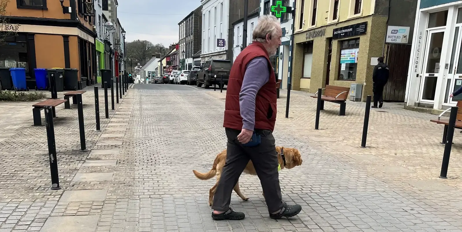 An older man in a maroon vest walks his golden dog through a quiet, pedestrian-friendly town street lined with colorful buildings and bollards.