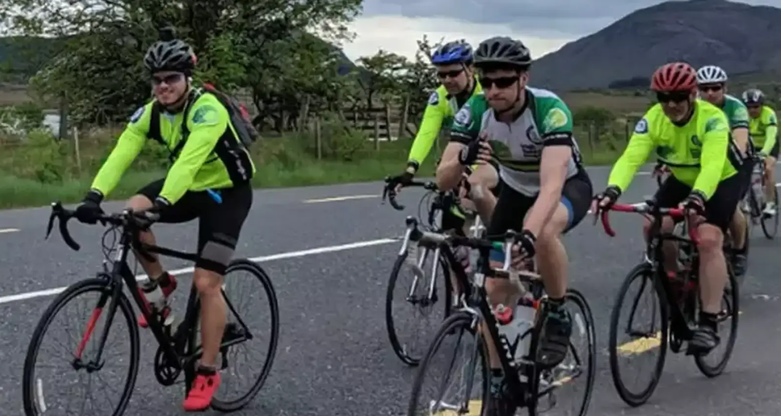 A group of cyclists wearing bright neon yellow and green jerseys ride together on a rural road with mountains visible in the background.