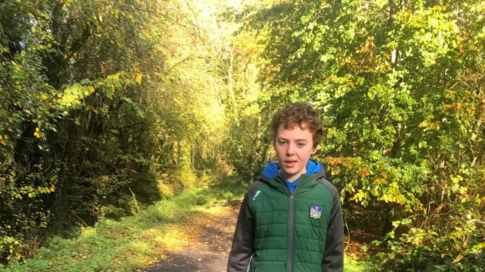 A young boy wearing a green quilted vest and blue hoodie stands on a tree-lined path dappled with golden sunlight filtering through the autumn foliage.