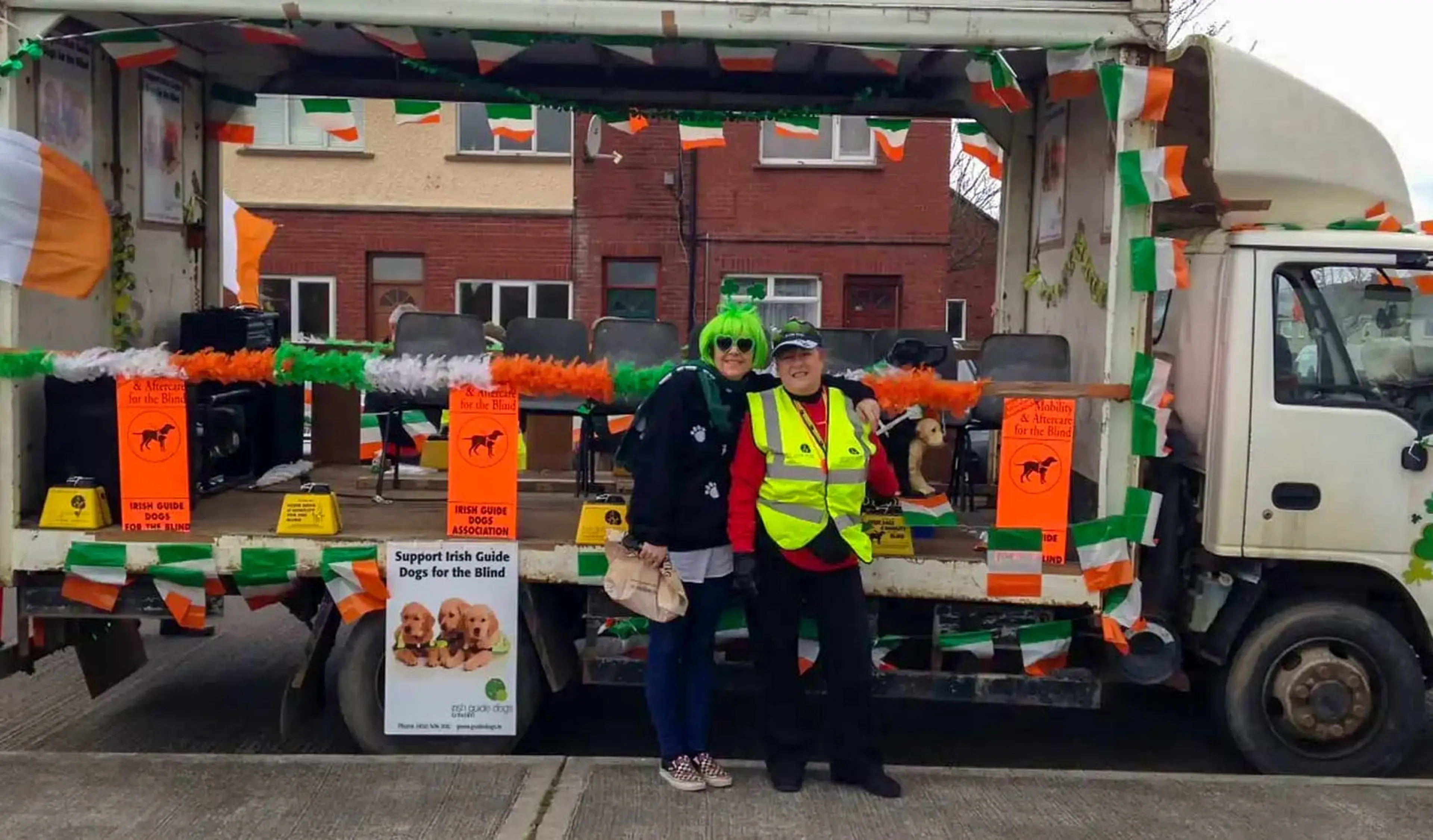 Two volunteers in festive attire stand in front of an Irish-decorated parade float promoting Irish Guide Dogs for the Blind, adorned with green, white, and orange flags and banners