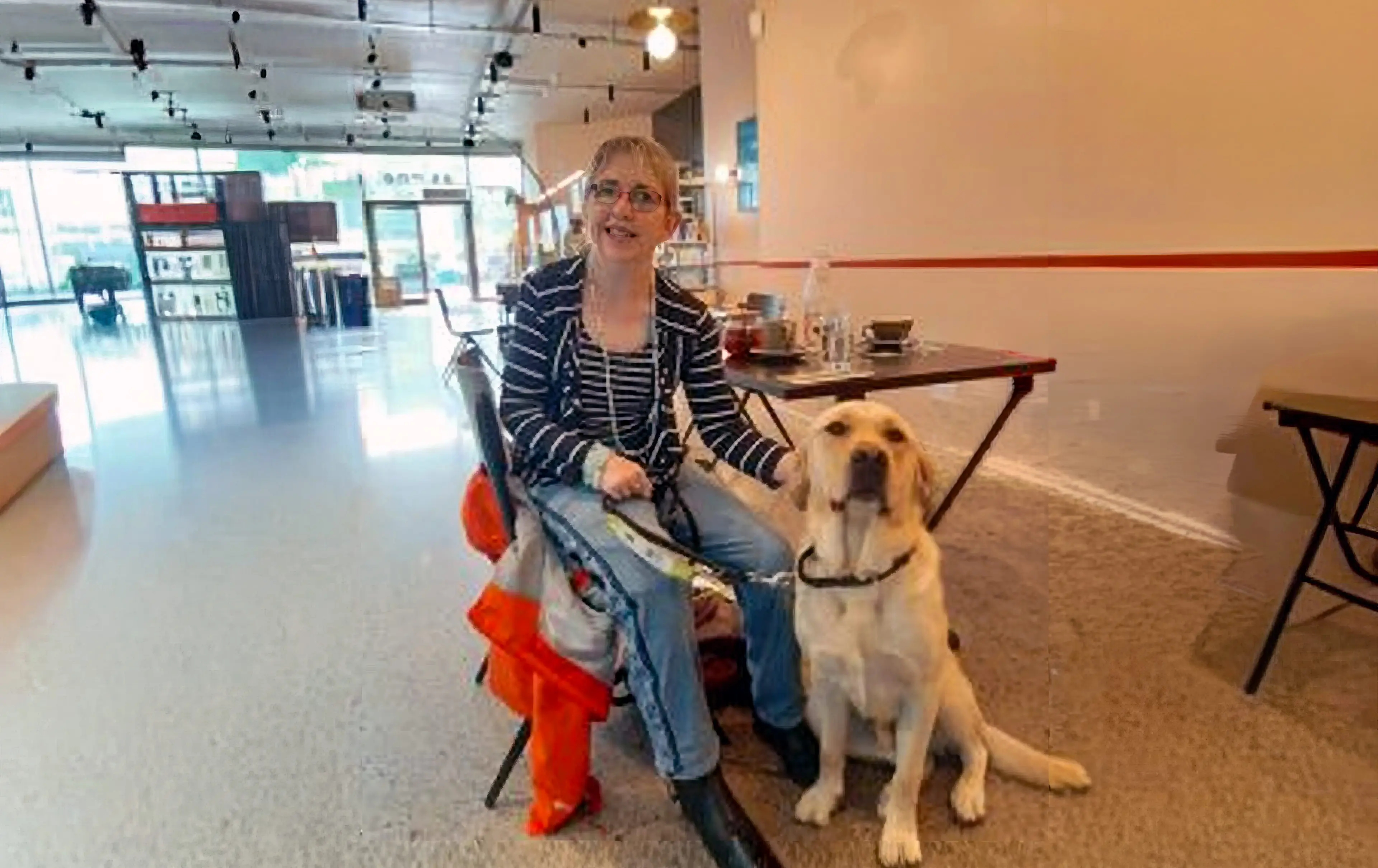 A woman wearing glasses and a striped blazer sits on a stool next to a yellow Labrador Retriever in a modern, well-lit indoor space.