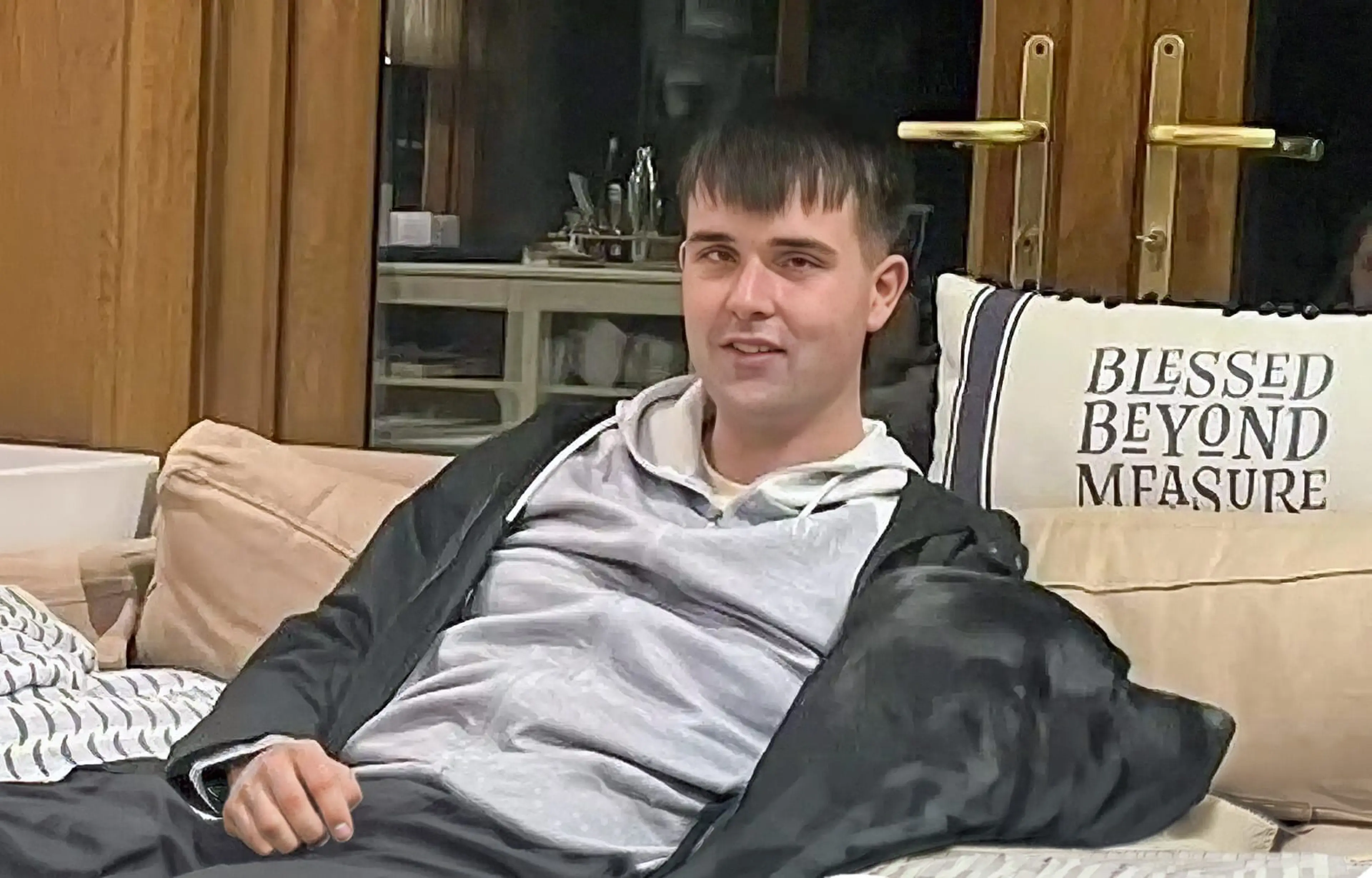A young man sits on a beige couch wearing a black jacket over a gray hoodie, with a pillow reading "Blessed Beyond Measure" visible behind him in a modern living room.
