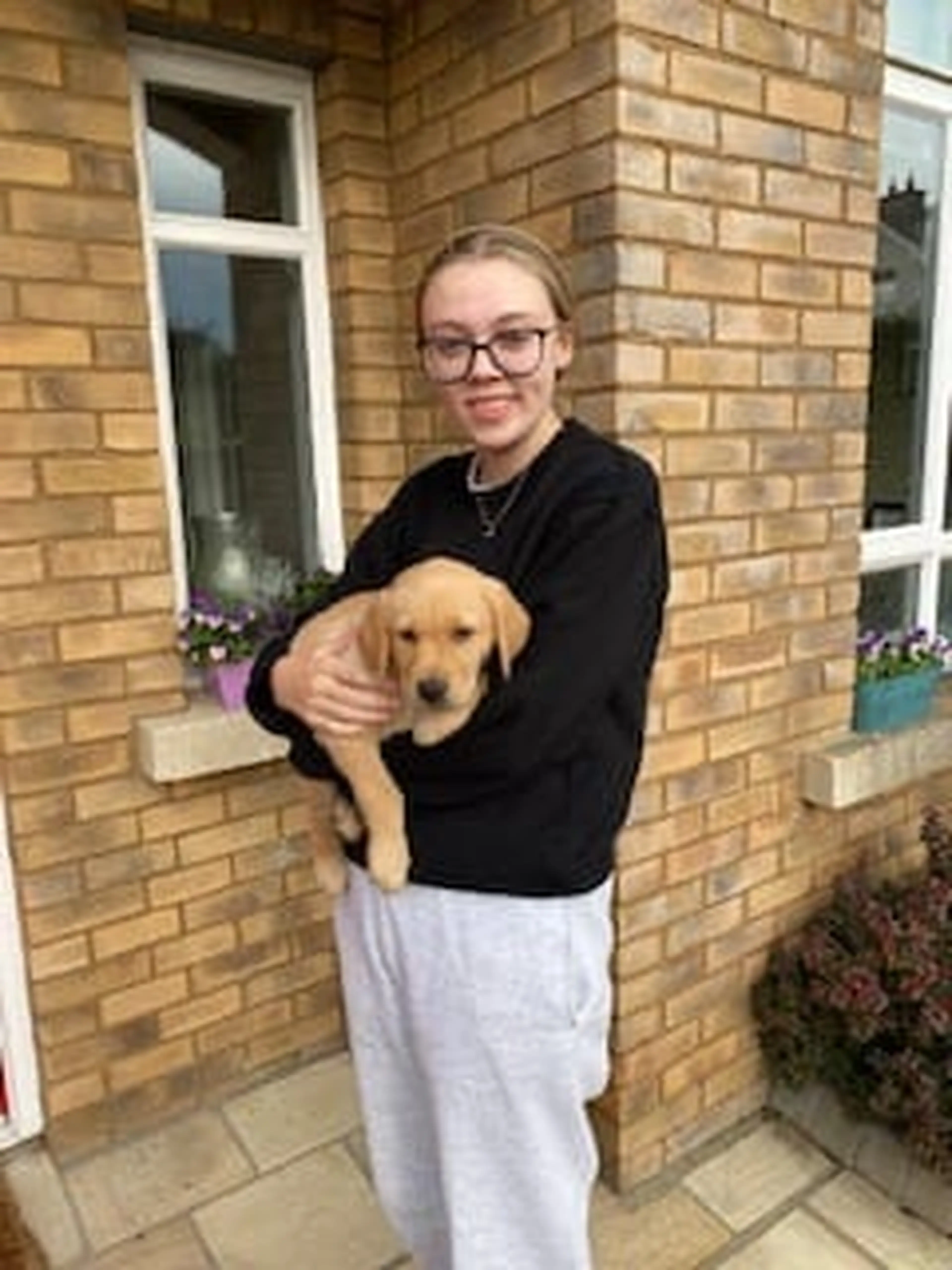 A person wearing glasses and dark clothing holds a yellow Labrador Retriever while standing in front of a brick house.