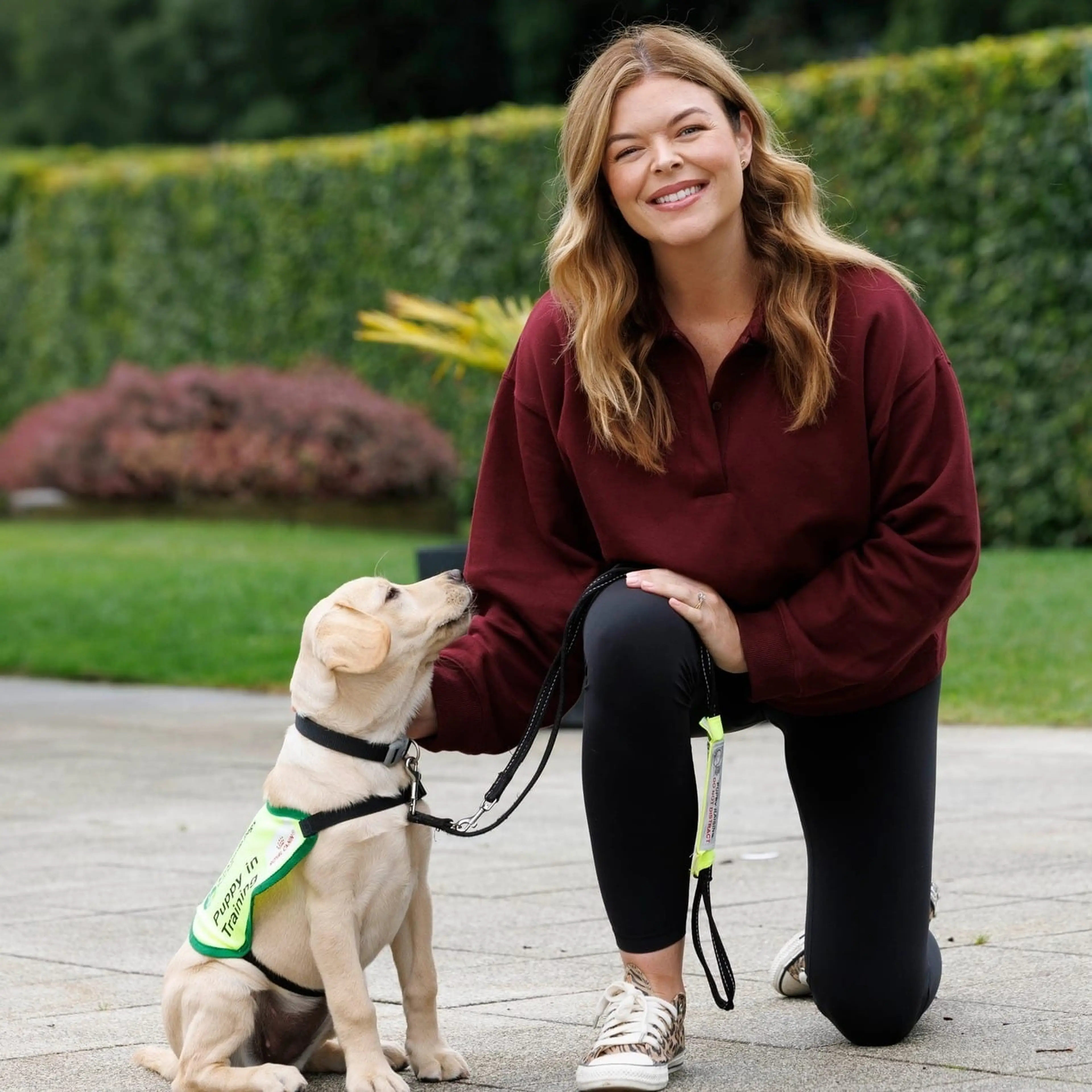 A woman in a maroon sweater crouches beside a yellow Labrador Retriever wearing a green service vest, smiling at the camera in a garden setting.