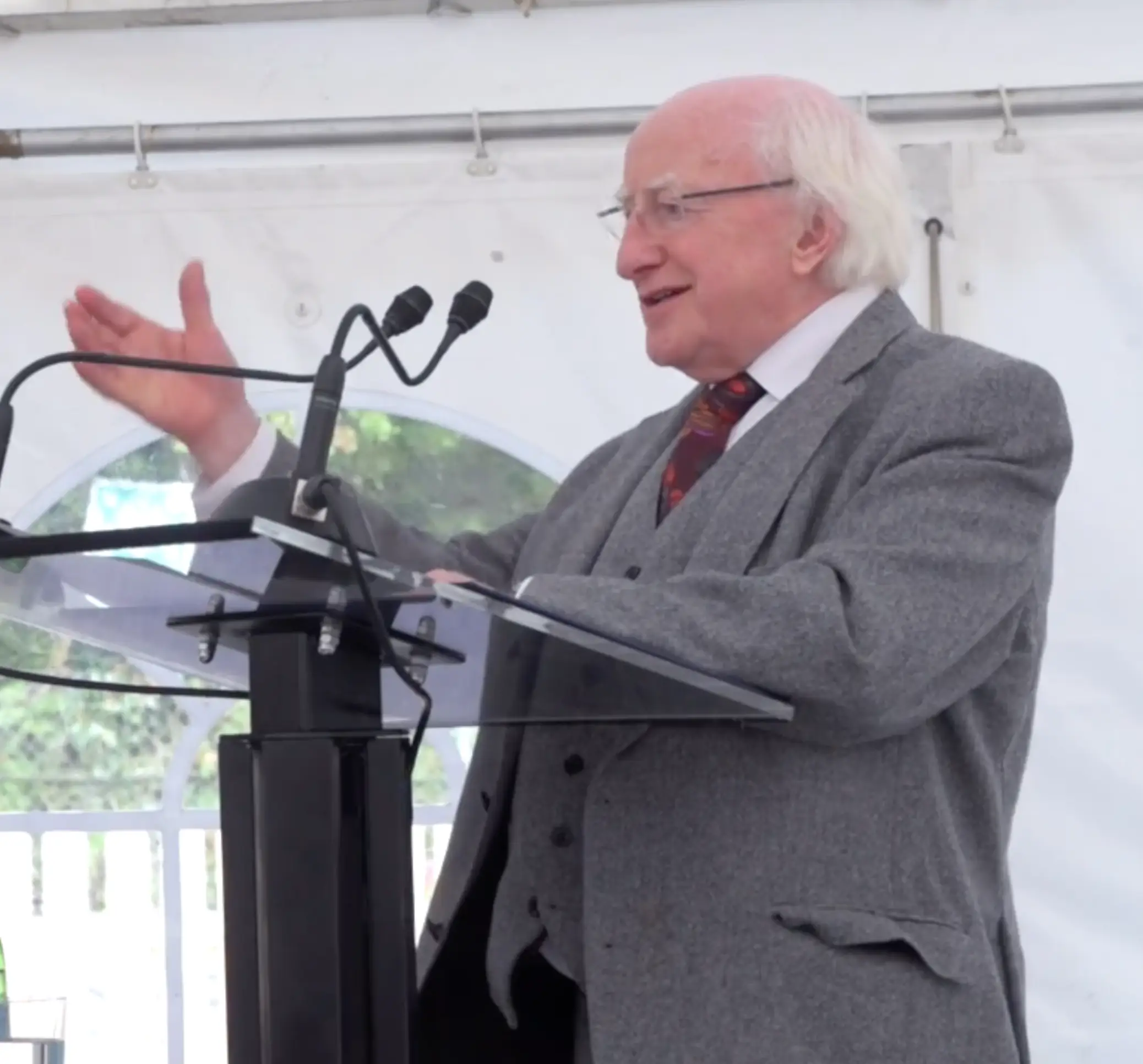 An older man with white hair and glasses, wearing a gray suit, standing at a podium and gesturing while speaking into microphones.
