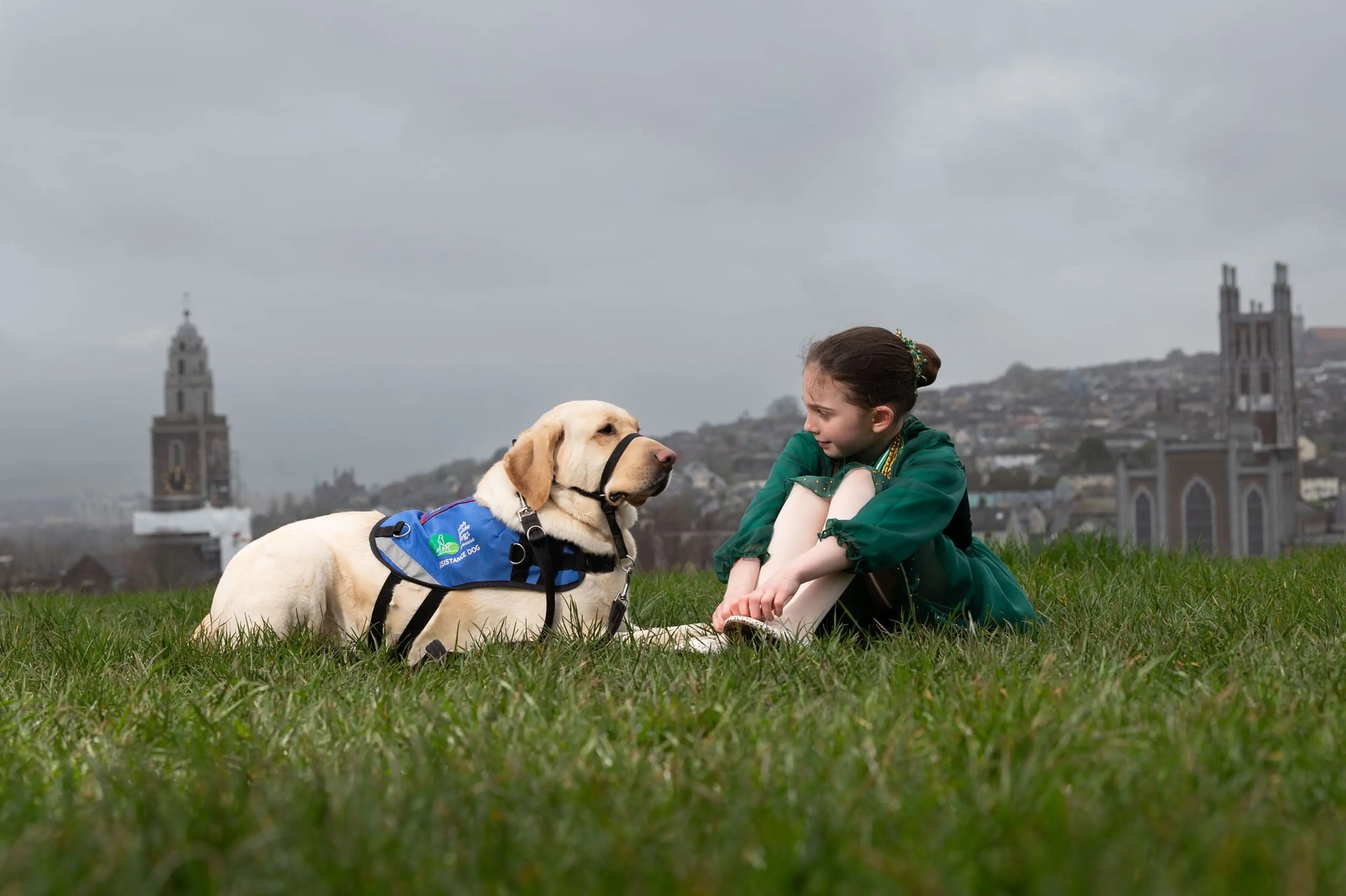 A young girl in a green jacket sits on grass next to a cream-colored service dog wearing a blue harness, with historic church spires visible in the overcast cityscape behind them.