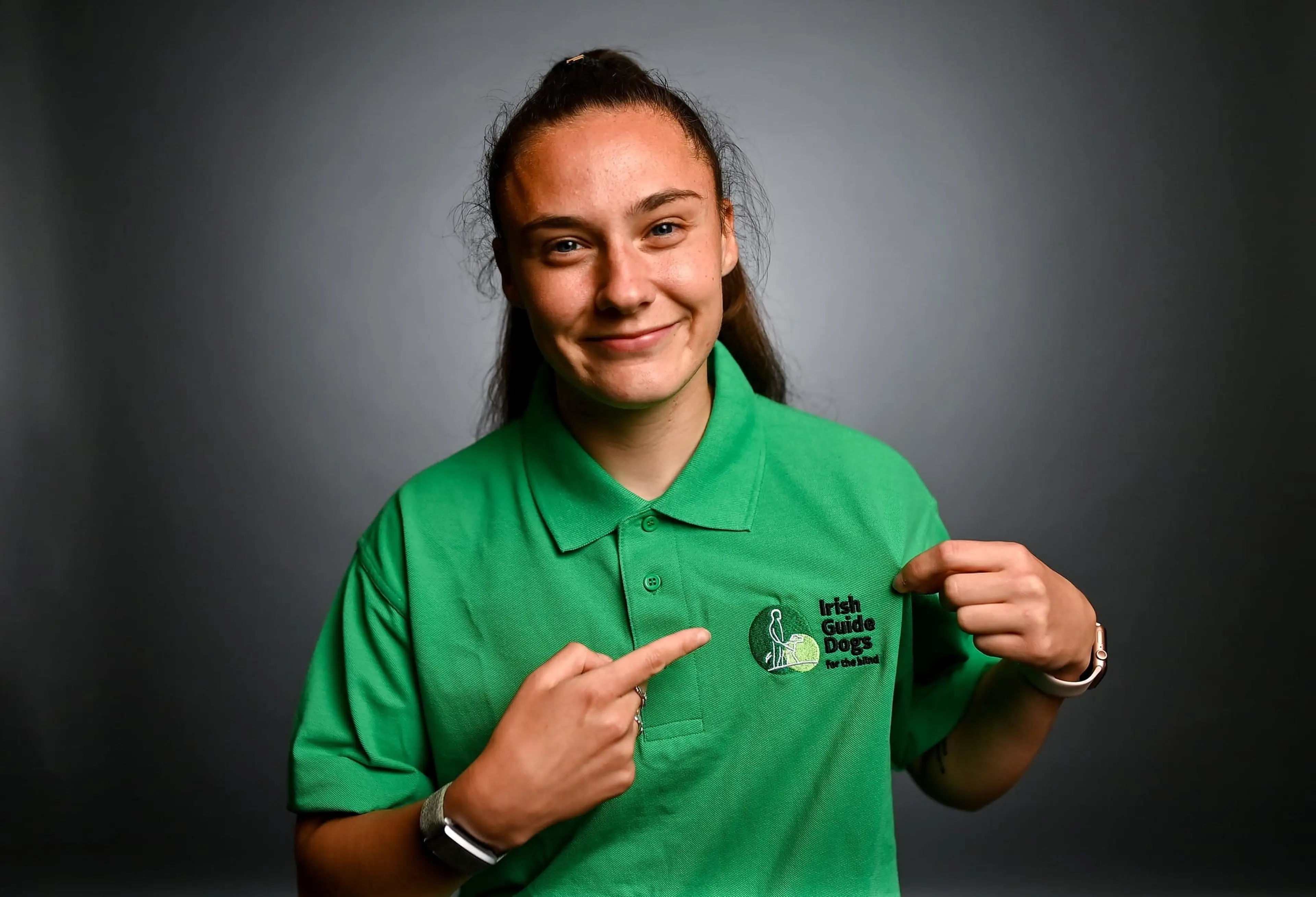 A young woman wearing a green Irish Guide Dogs polo shirt points to the organization's logo on her chest while smiling at the camera against a gray background.