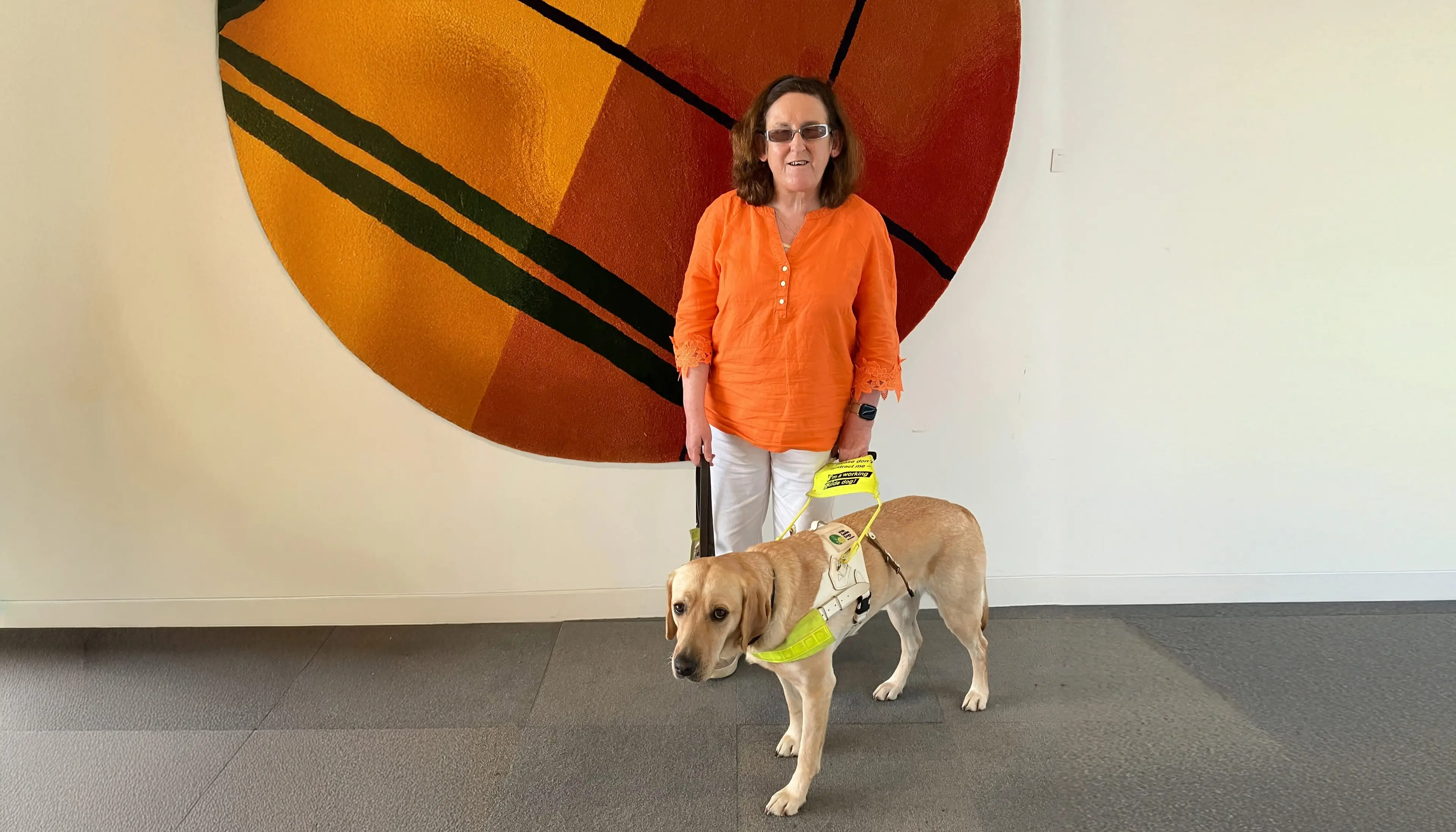 A woman wearing an orange top and sunglasses stands beside a yellow Labrador service dog wearing a harness and leash against a colorful abstract artwork.